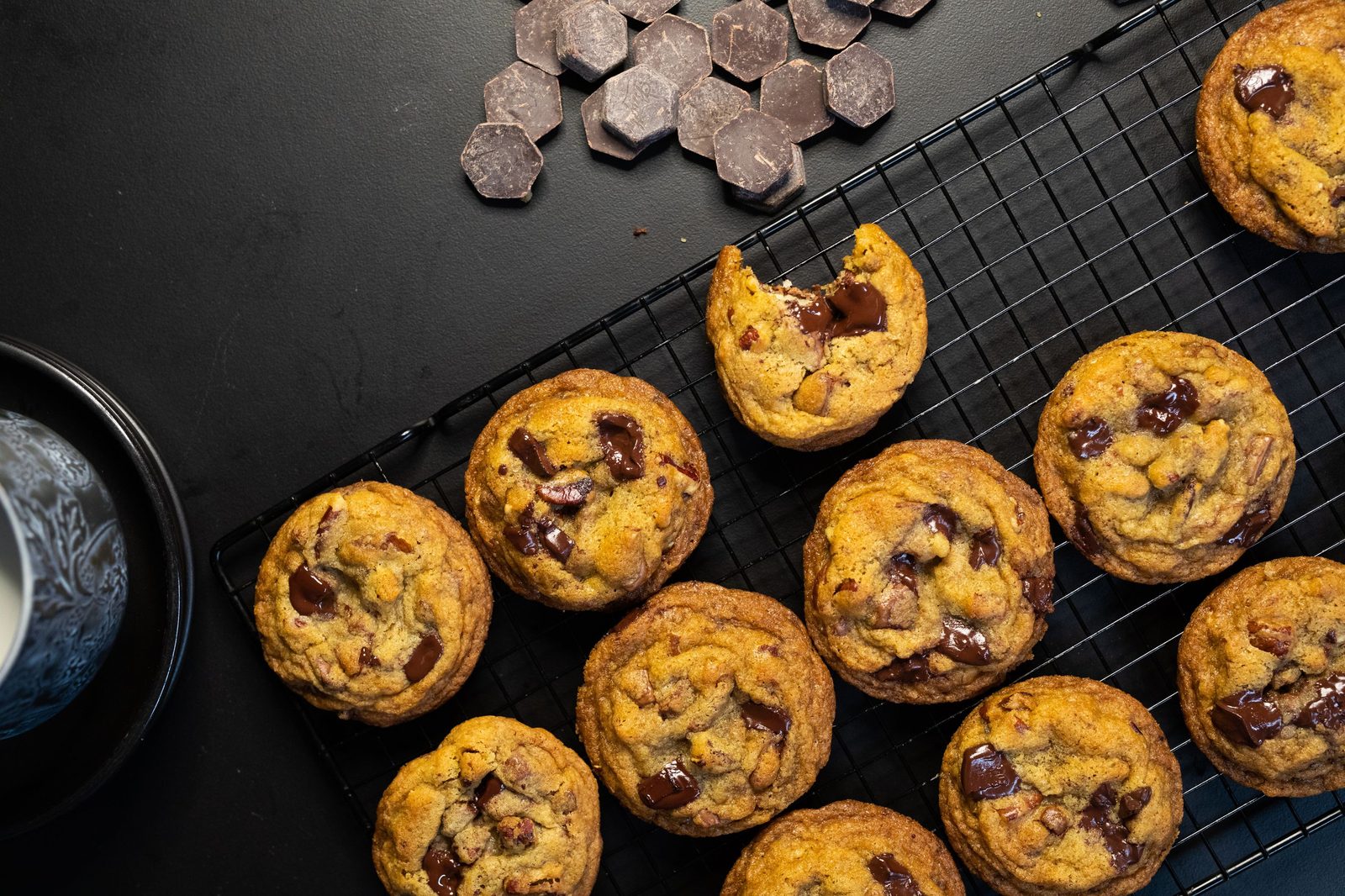 Chocolate chip cookies on a cooling rack, one bitten, beside a pile of milk chocolate chunks, from above