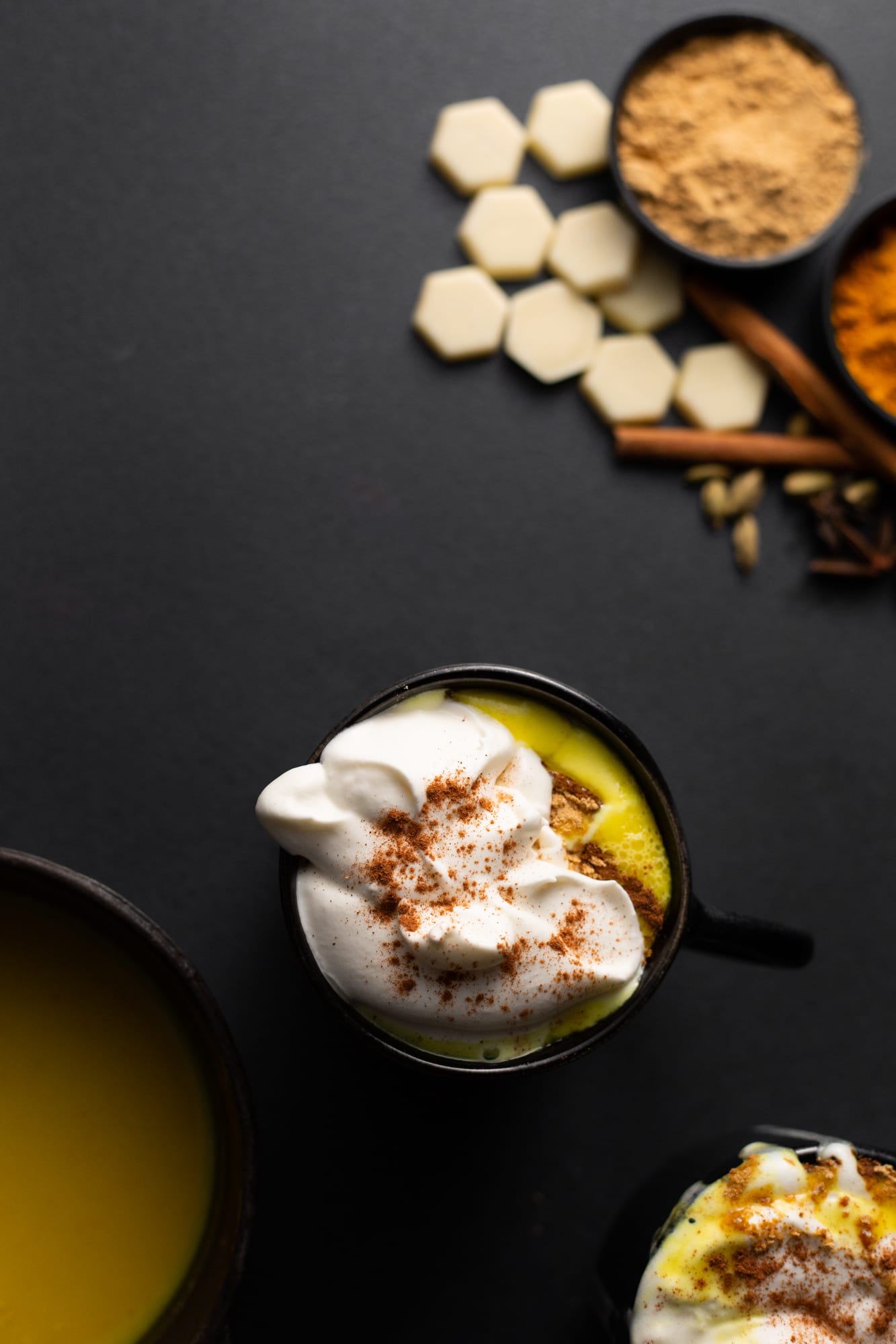 Mugs of turmeric and ginger white hot chocolate topped with cream, with spices and white chocolate, from above