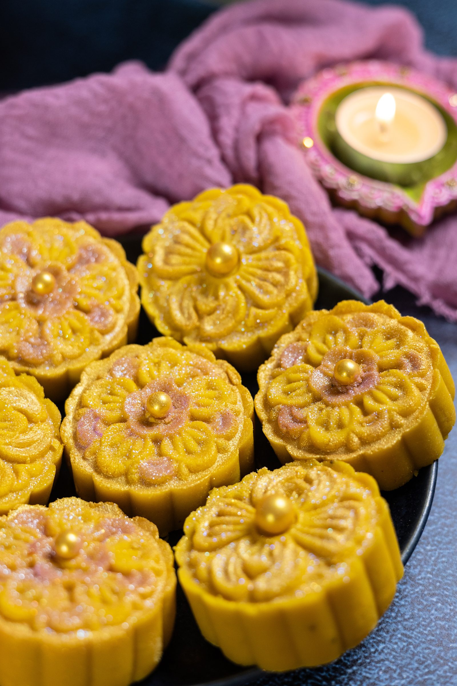 Mango burfee flowers on a plate at an angle, with a pink cloth and lit diya behind
