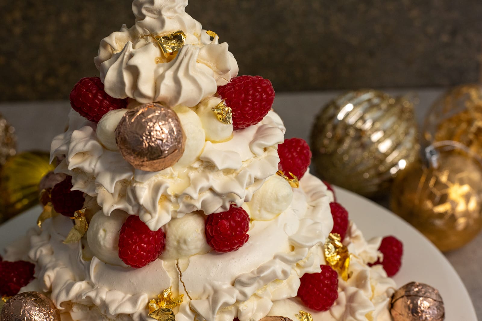 The tiers of a Christmas tree pavlova with raspberries and gold baubles around it