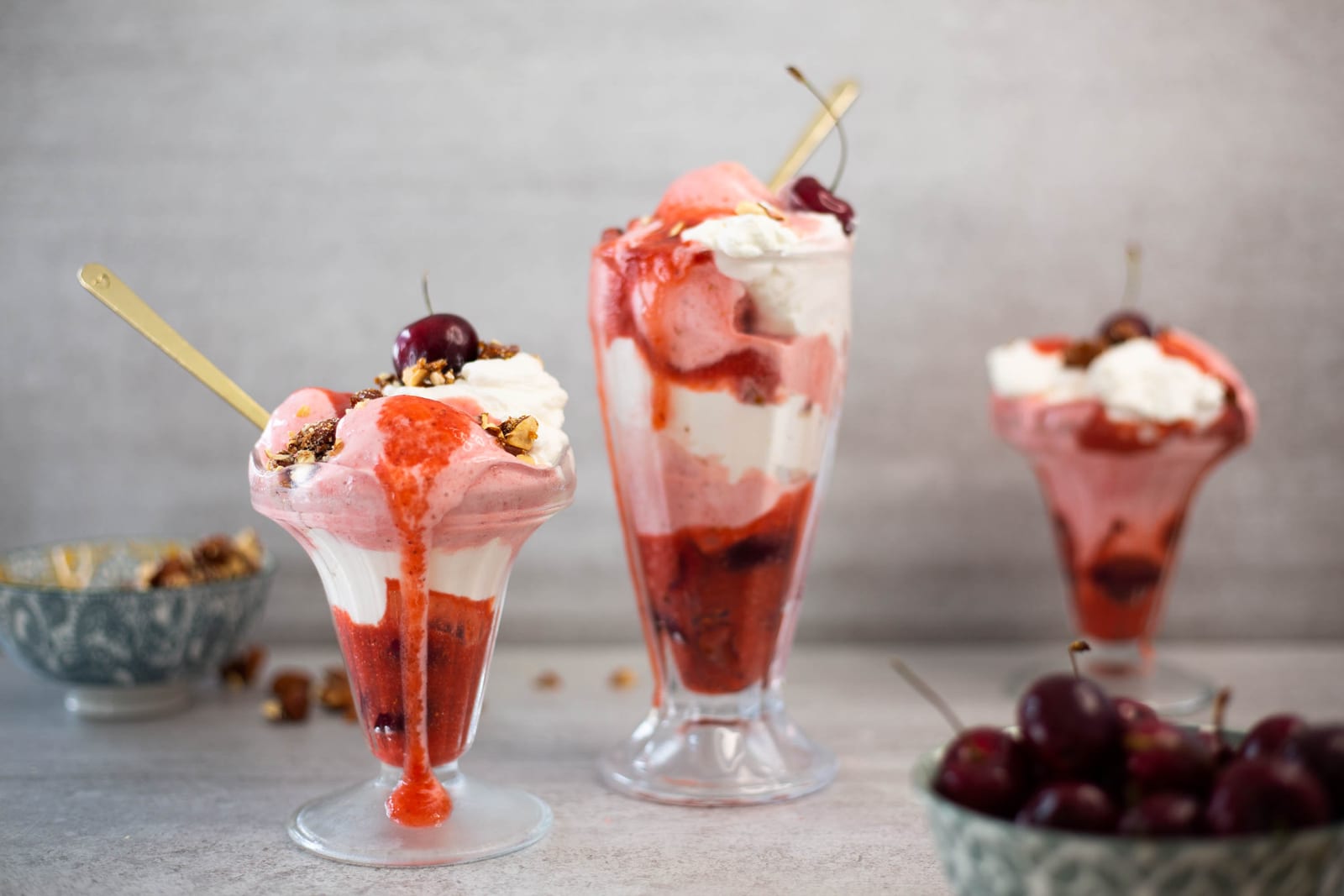 Three berries and cream sundaes in a row, raspberry sauce dripping down the glasses beside a bowl of cherries