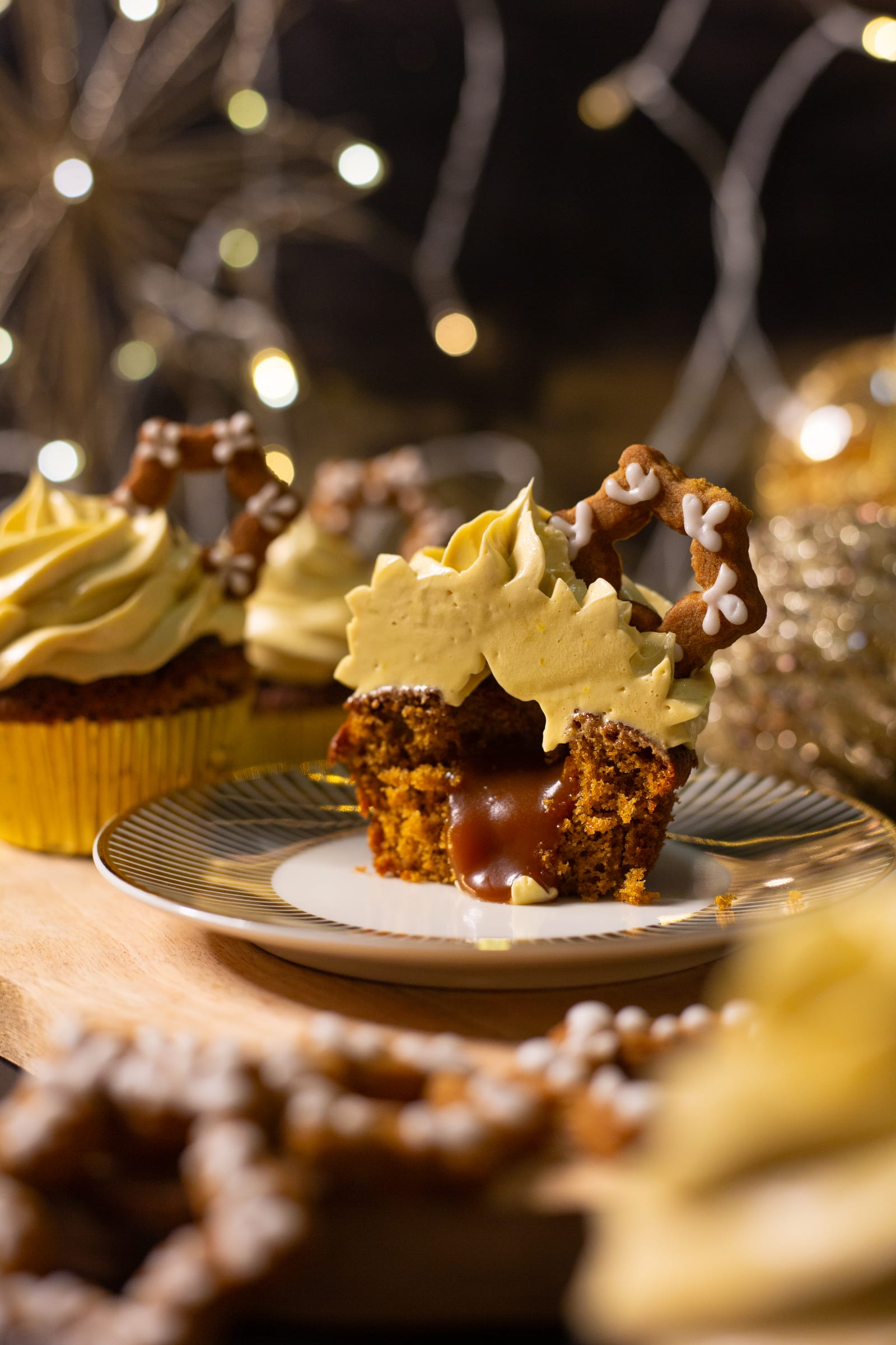 Gingerbread cupcakes topped with pretzels, a cut one oozing caramel in front, against fairy lights