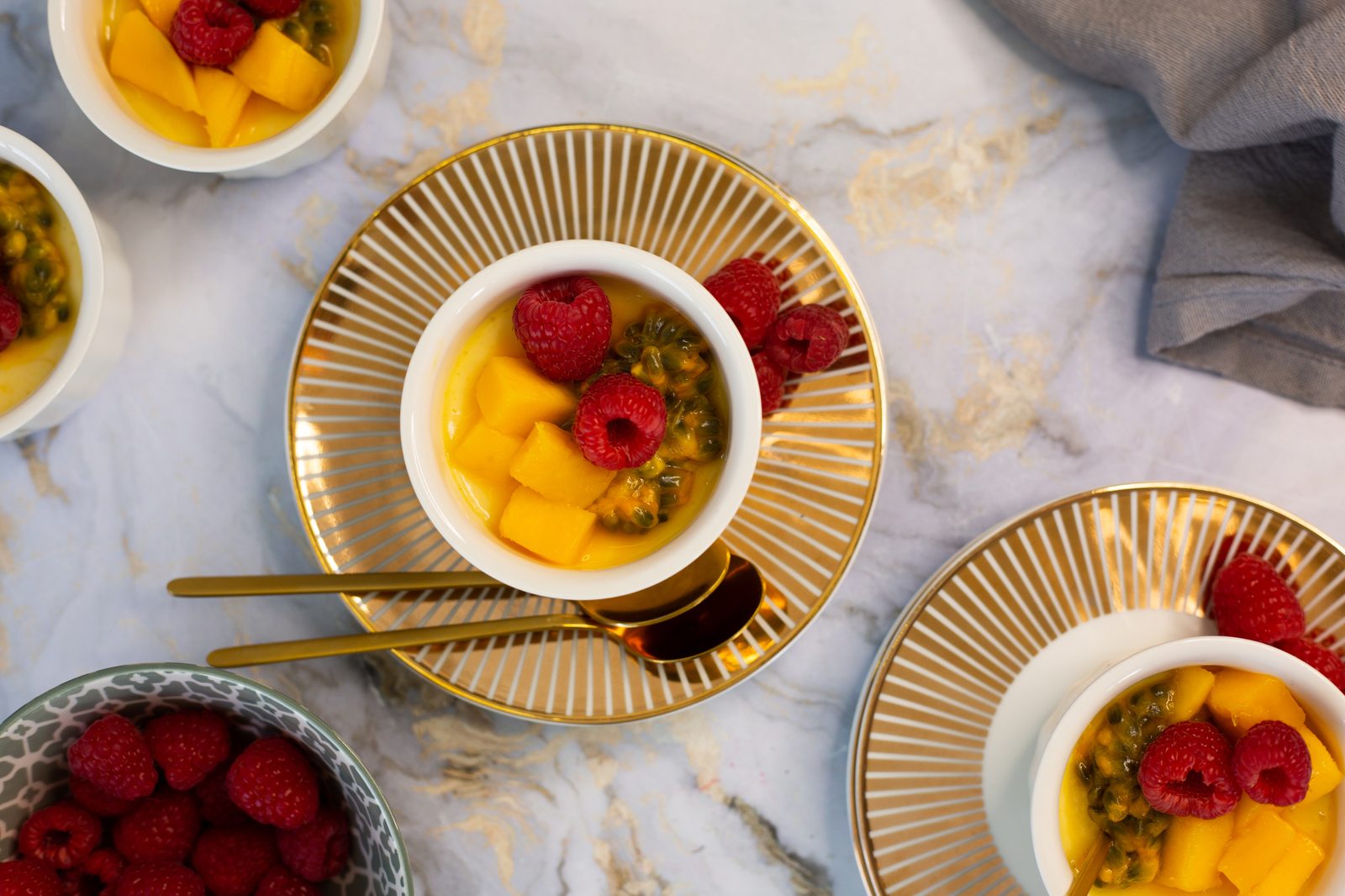 A coconut, mango and granadilla baked yogurt on a gold plate from above, among more ramekins and a bowl of raspberries