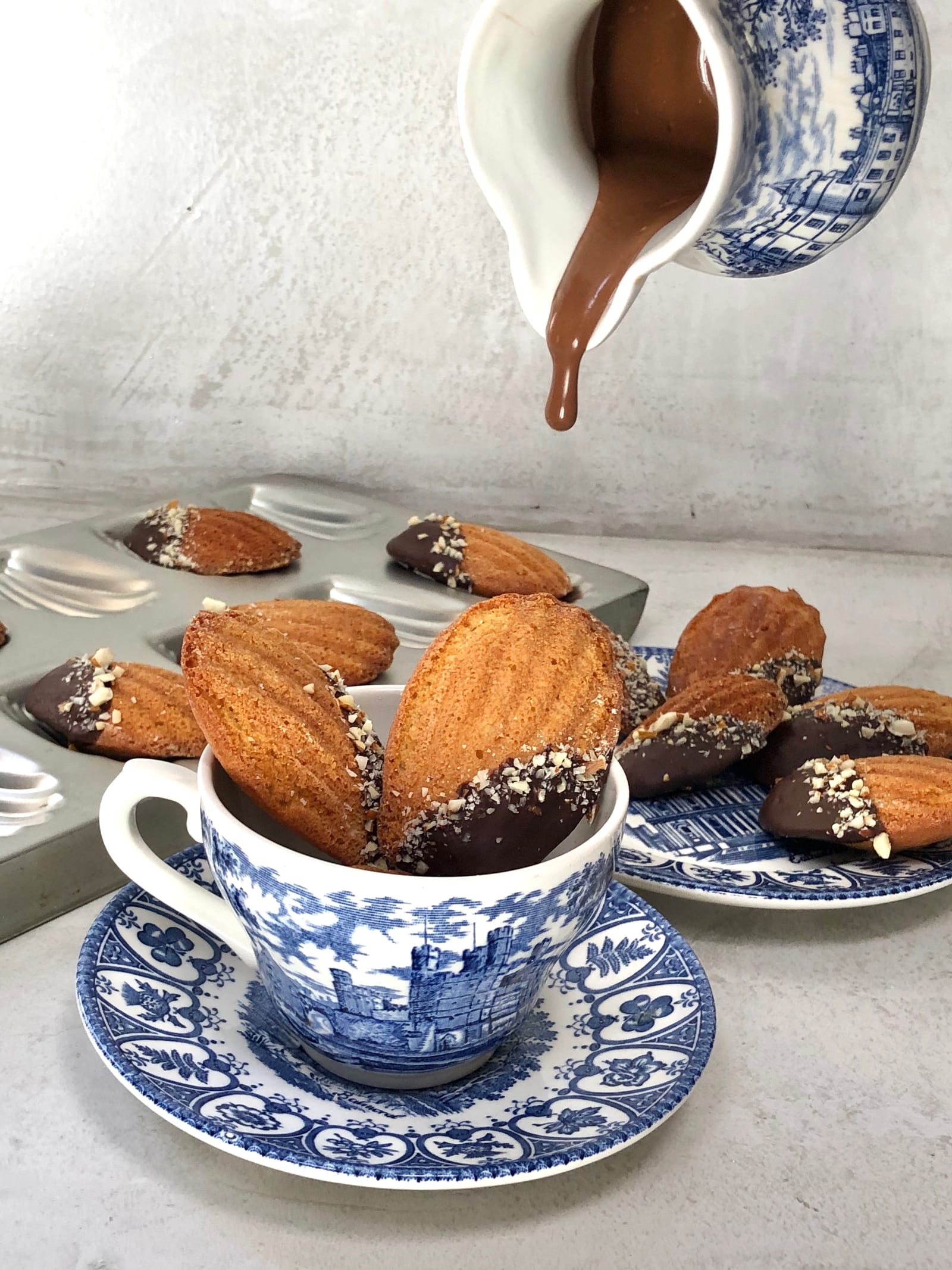 Chocolate being poured from a jug over a cup heaped with brown butter and almond madeleines