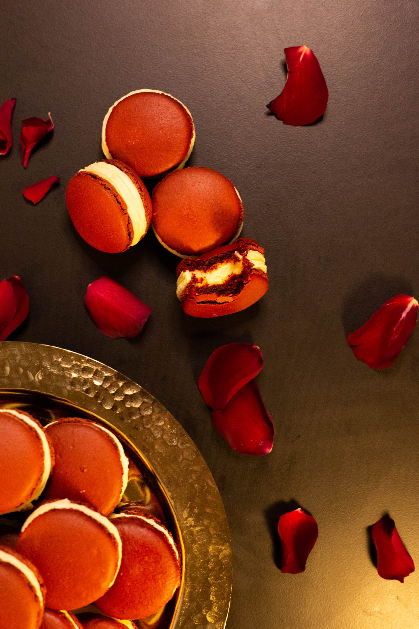 Red velvet macarons and a bitten one beside a gold bowl and rose petals, from above