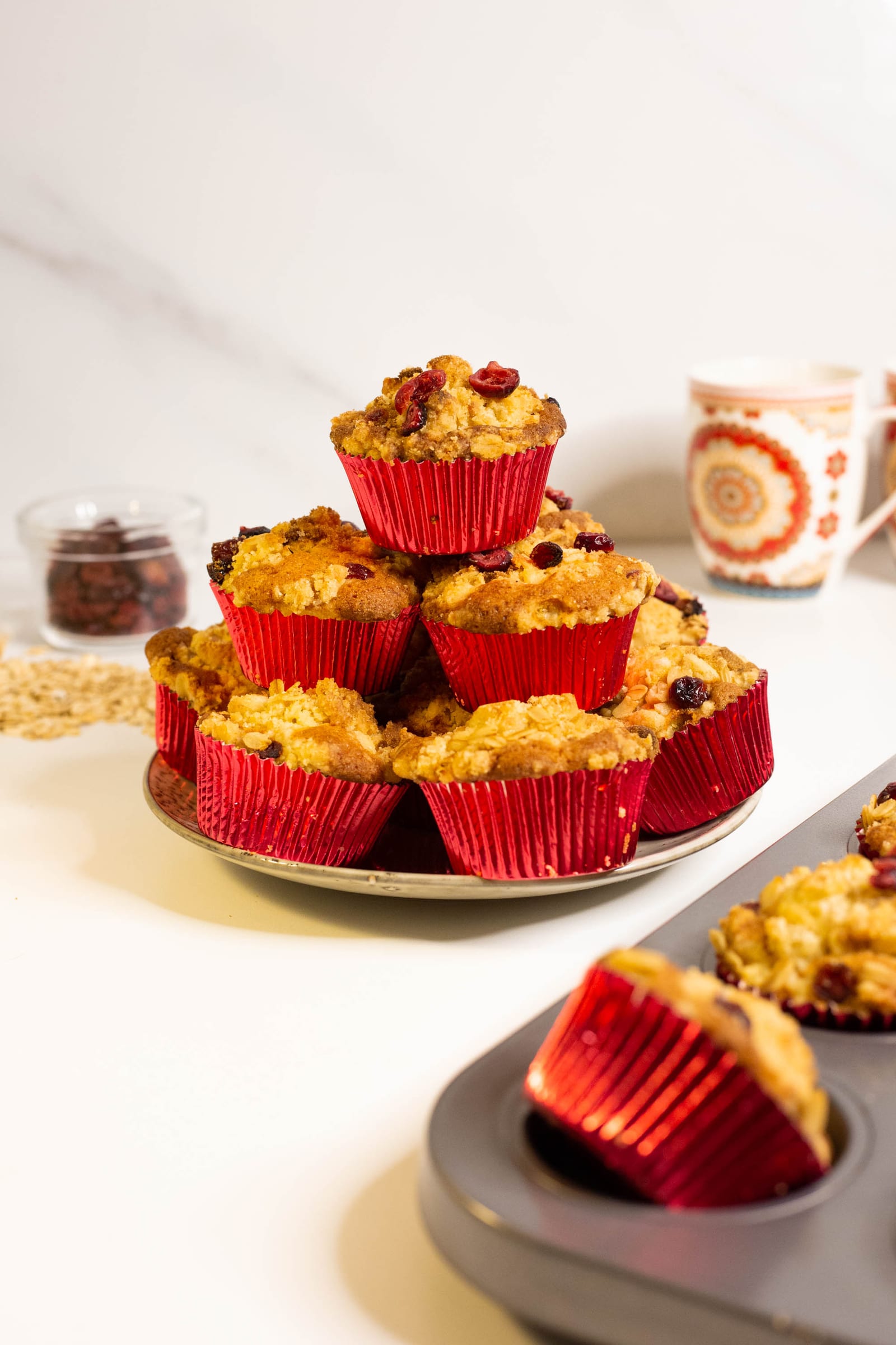 A pile of cranberry crumble muffins in red cases on a plate, the muffin tin in front