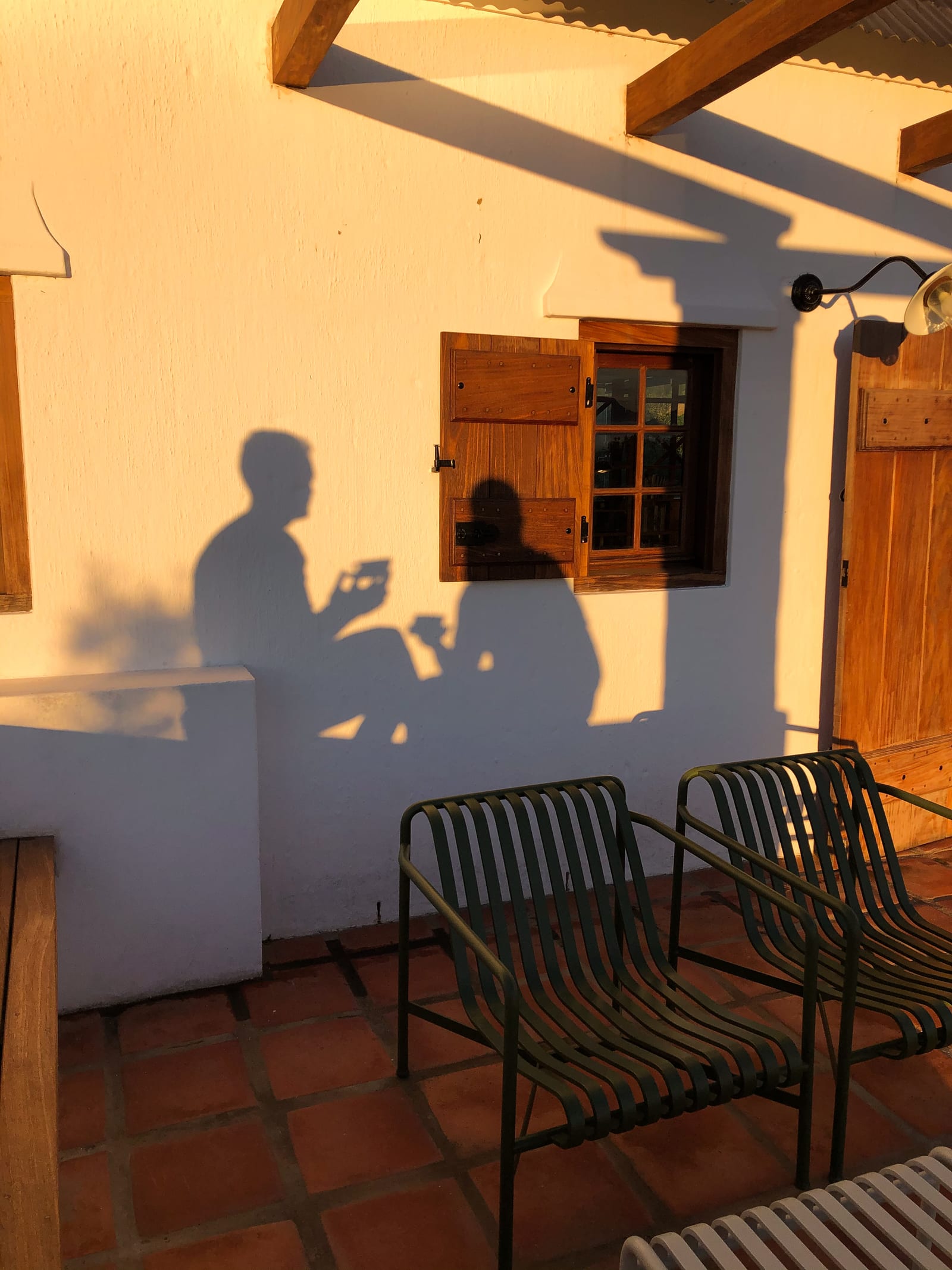 The shadows of two people with drinks on a cottage stoep, cast on a whitewashed wall beside a shuttered window