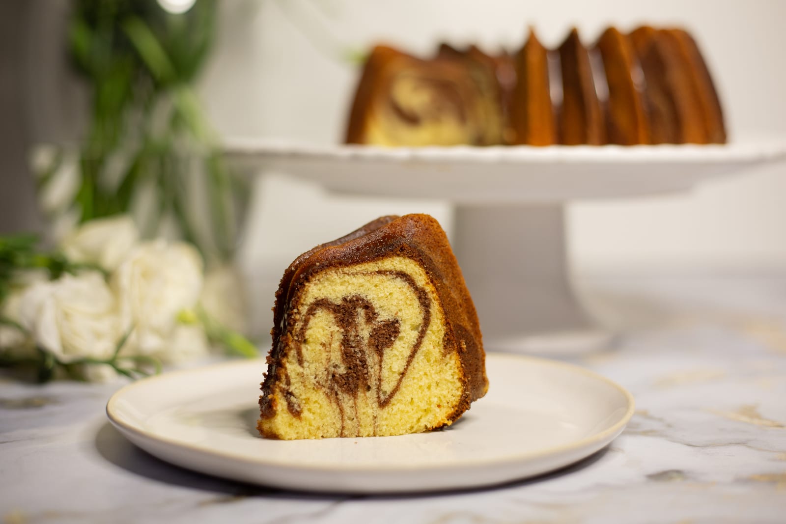 A slice of marble pound cake showing its chocolate swirl, with the rest of the bundt on a cake stand behind