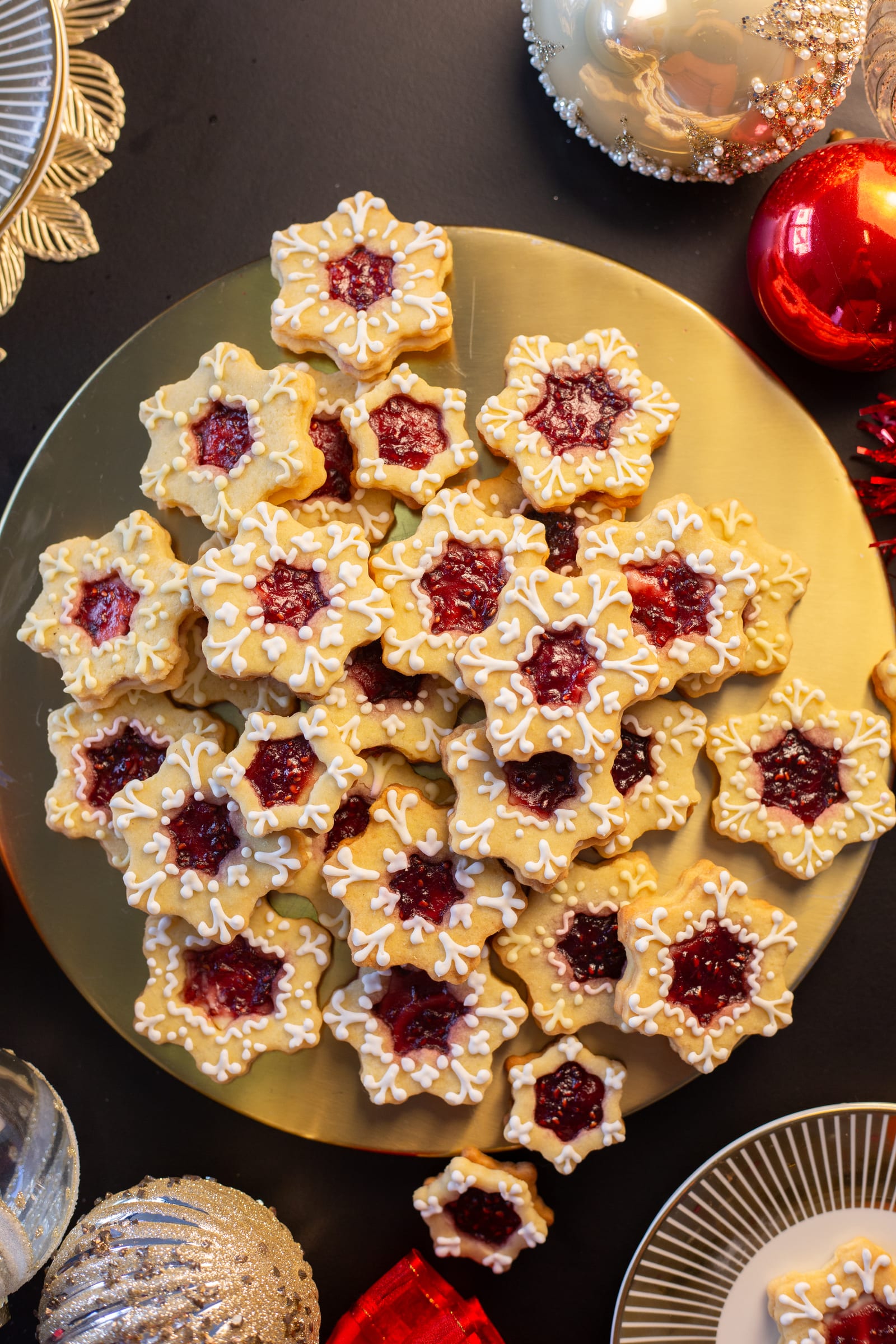 A gold platter piled with star-shaped lemon and raspberry Linzer cookies, surrounded by Christmas baubles
