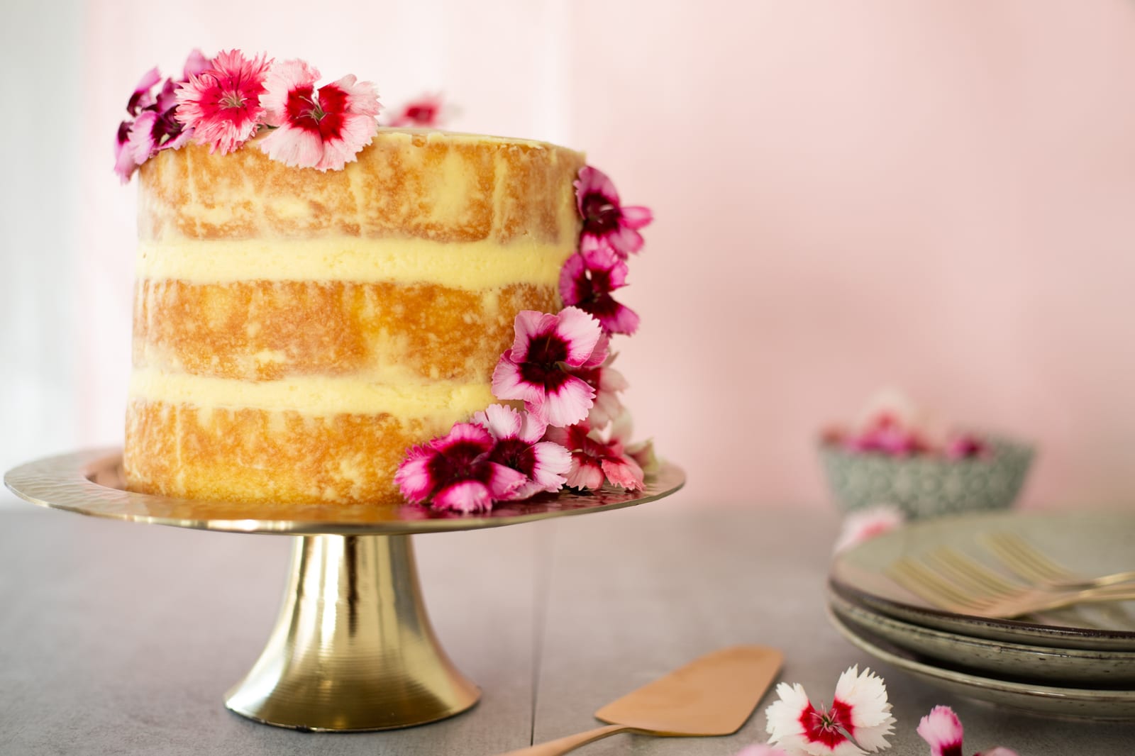 A lemon curd cake with pink flowers cascading down it on a gold stand, plates and a cake server beside it