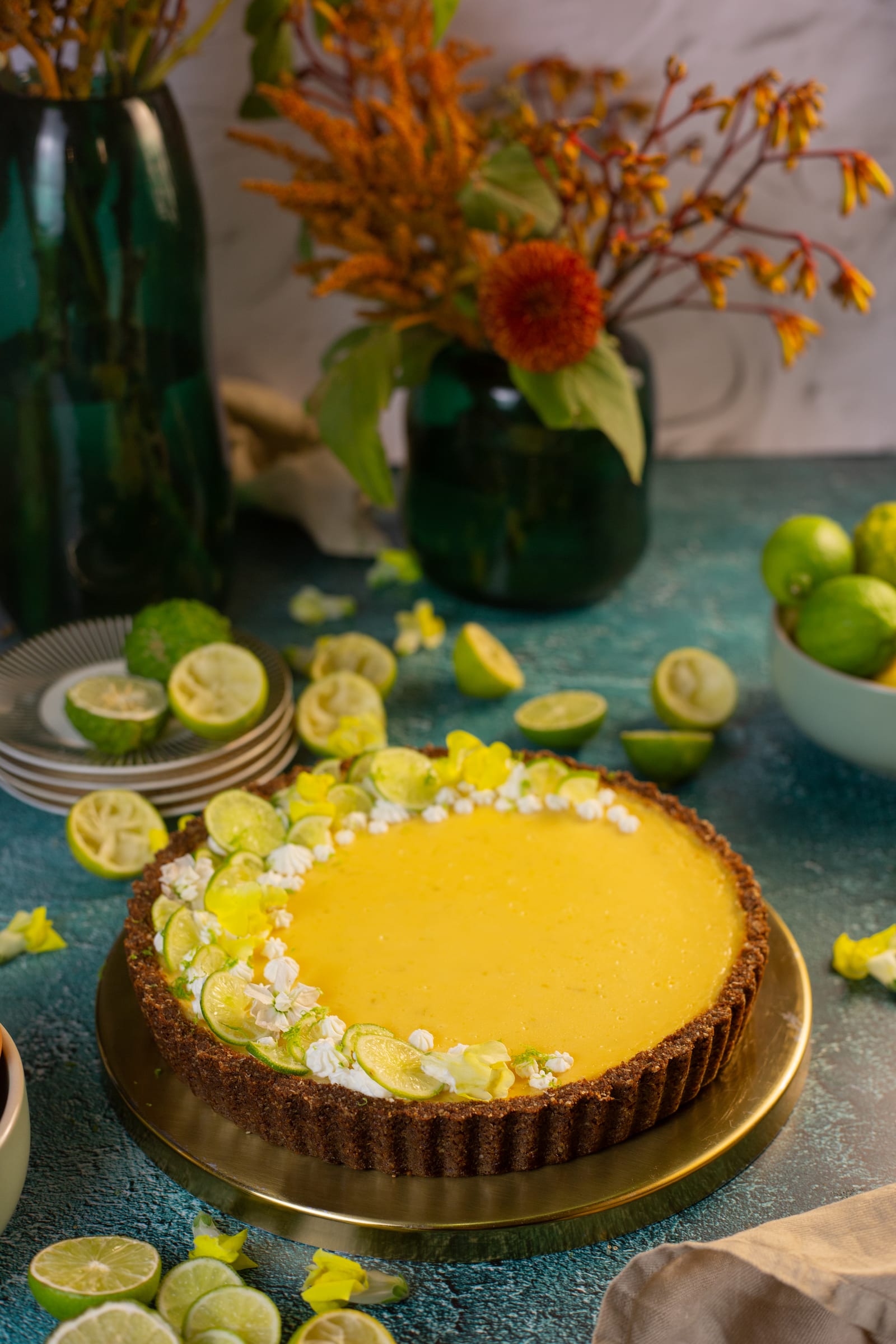 The key lime pie on a gold plate, with a vase of flowers and limes behind