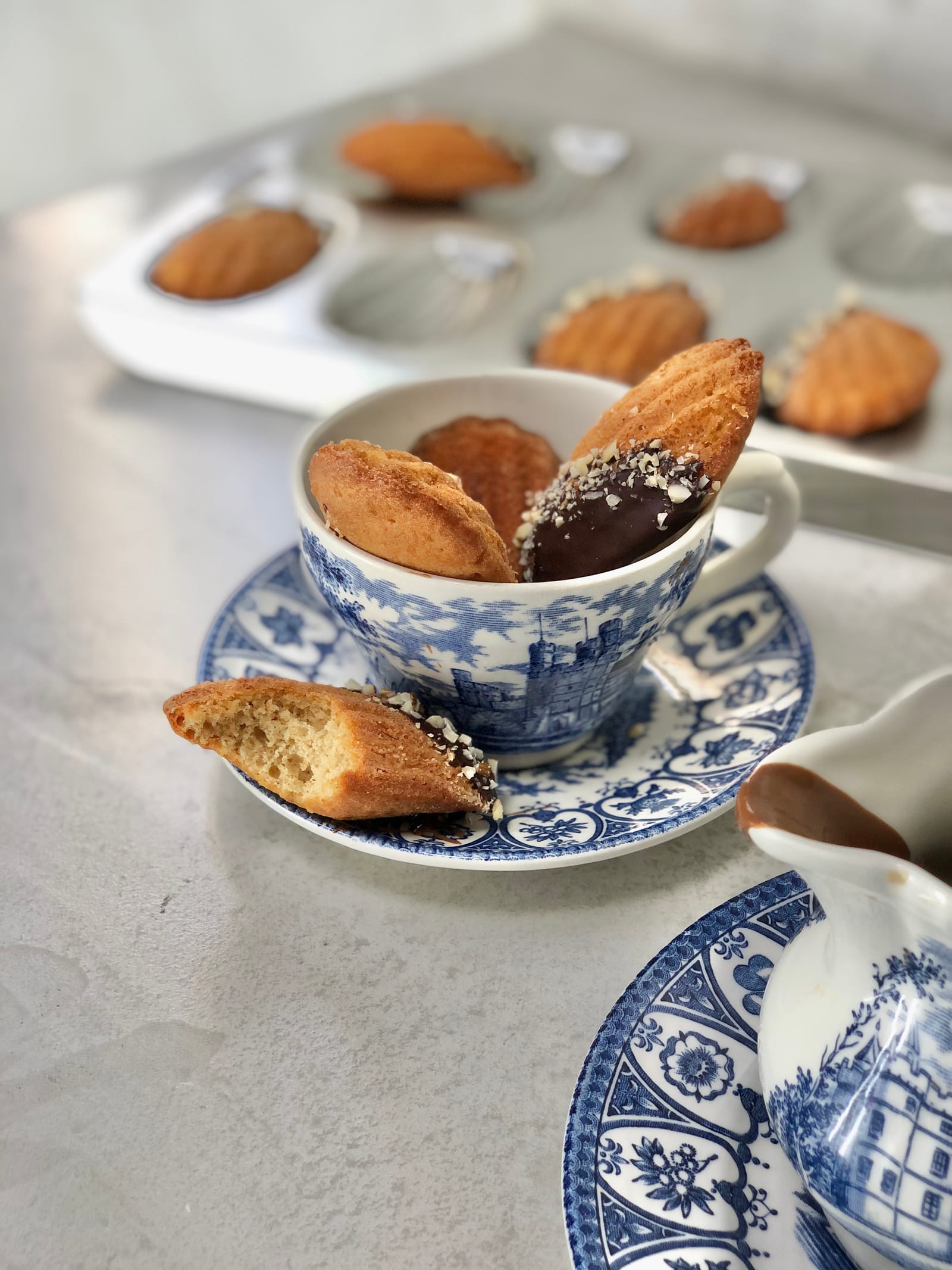 A broken brown butter and almond madeleine showing its crumb, a cup of madeleines behind