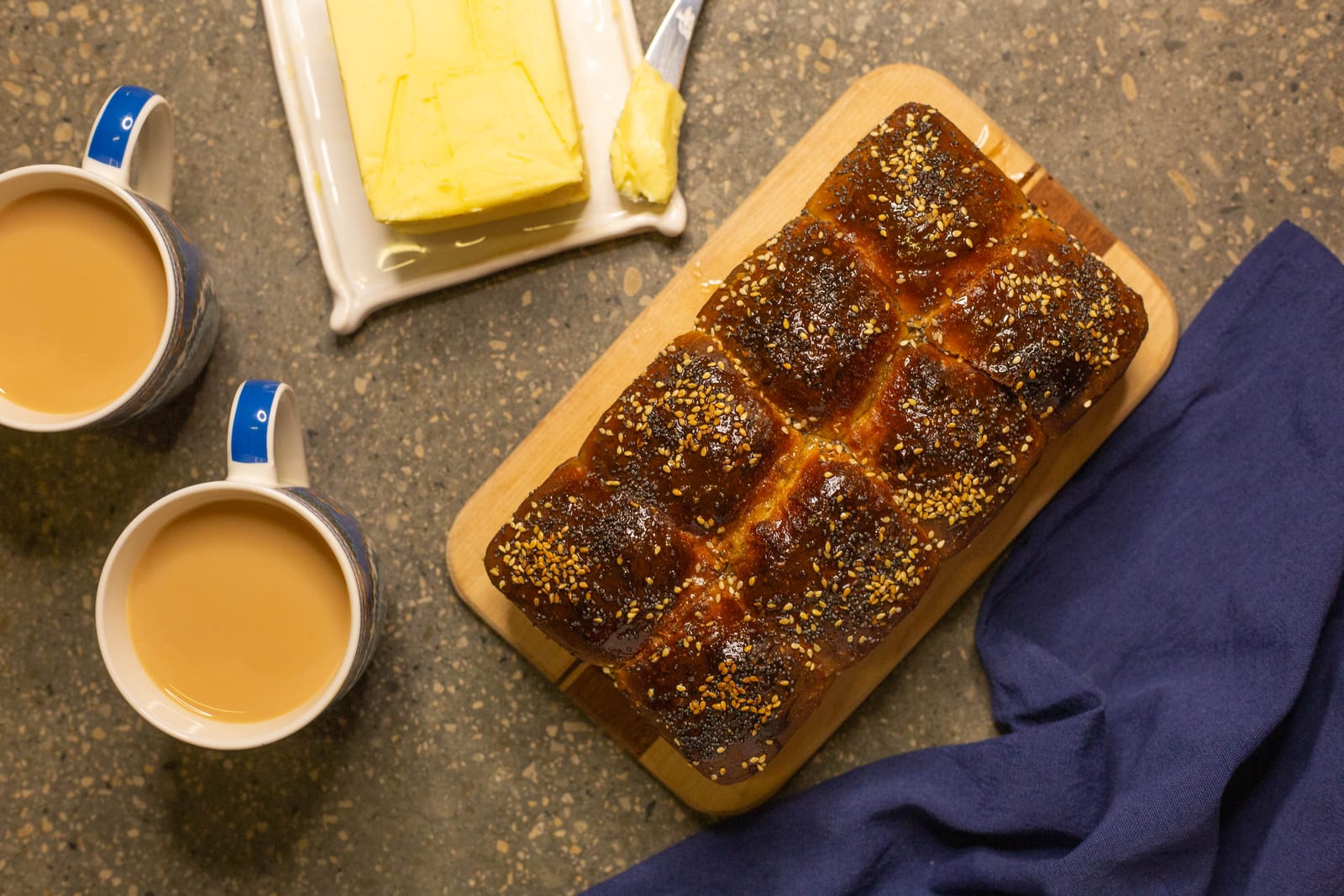A seeded mosbolletjies loaf on a wooden board beside two cups of milky tea and a block of butter