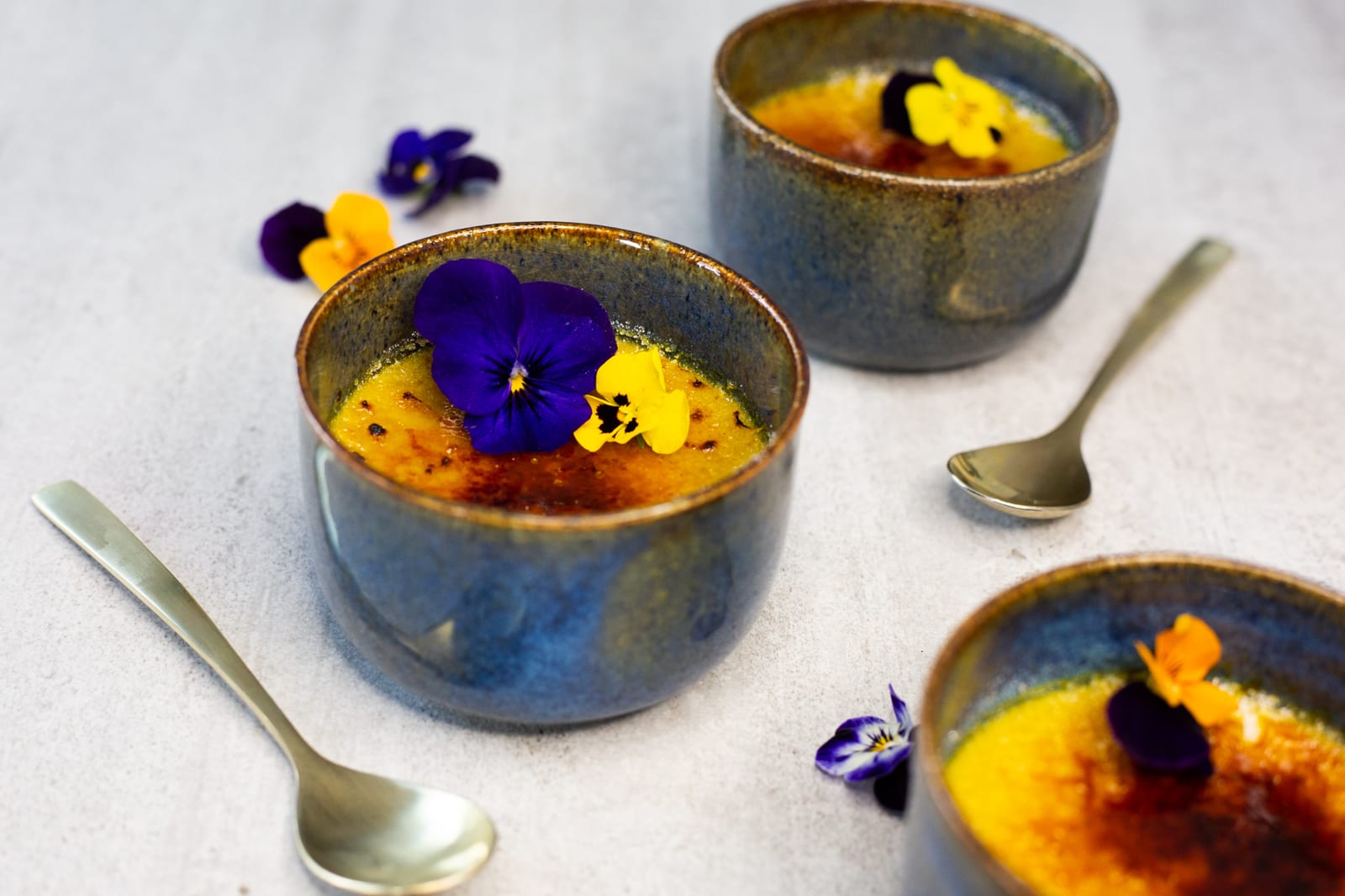 Three chai-spiced crème brûlées with violas and gold spoons