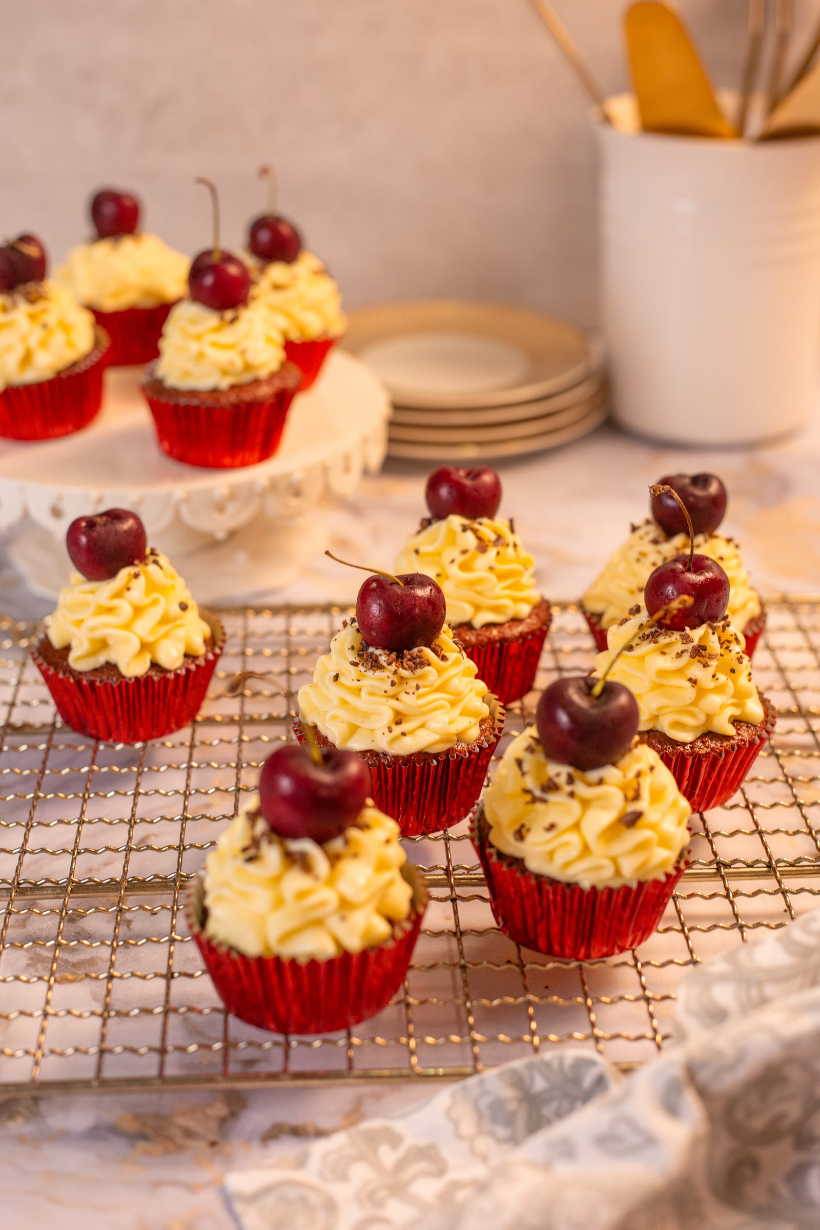 Red velvet cupcakes with cherries on a cooling rack, with plates behind
