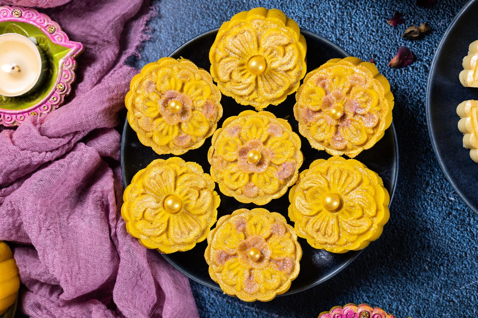 A dark plate of flower-shaped mango burfee with gold pearls, from above, beside a lit diya