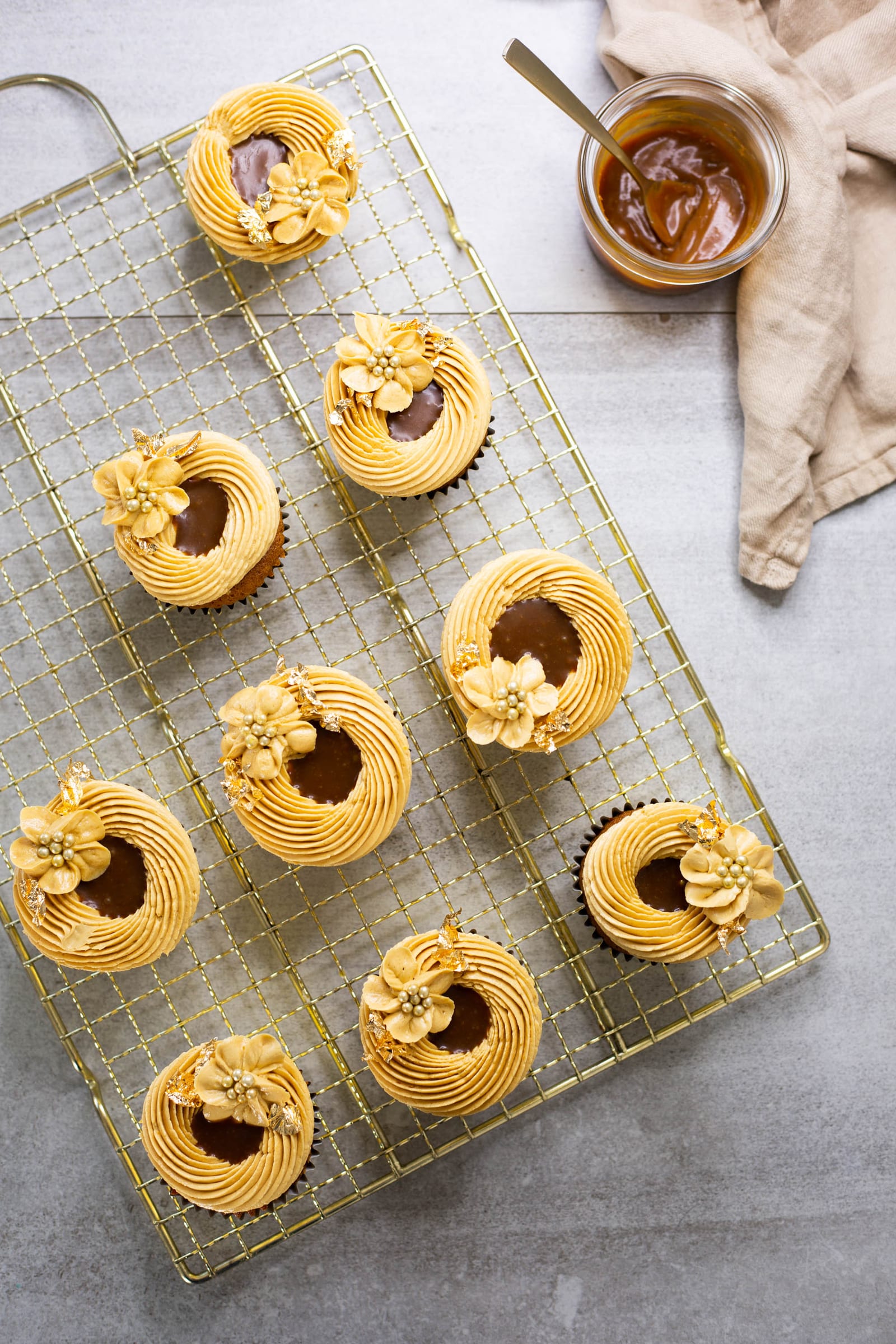 A batch of salted caramel cupcakes on a gold wire rack from above, with a jar of caramel sauce