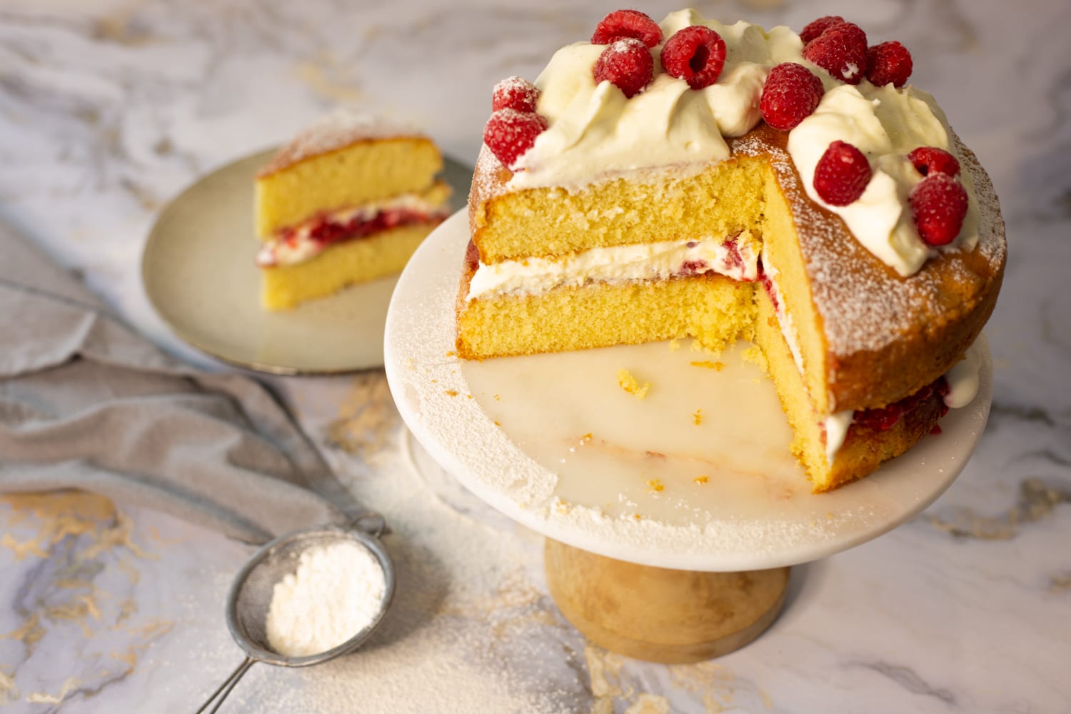 A Victoria sandwich with a wedge cut out on a marble cake stand, a slice on a plate beside it