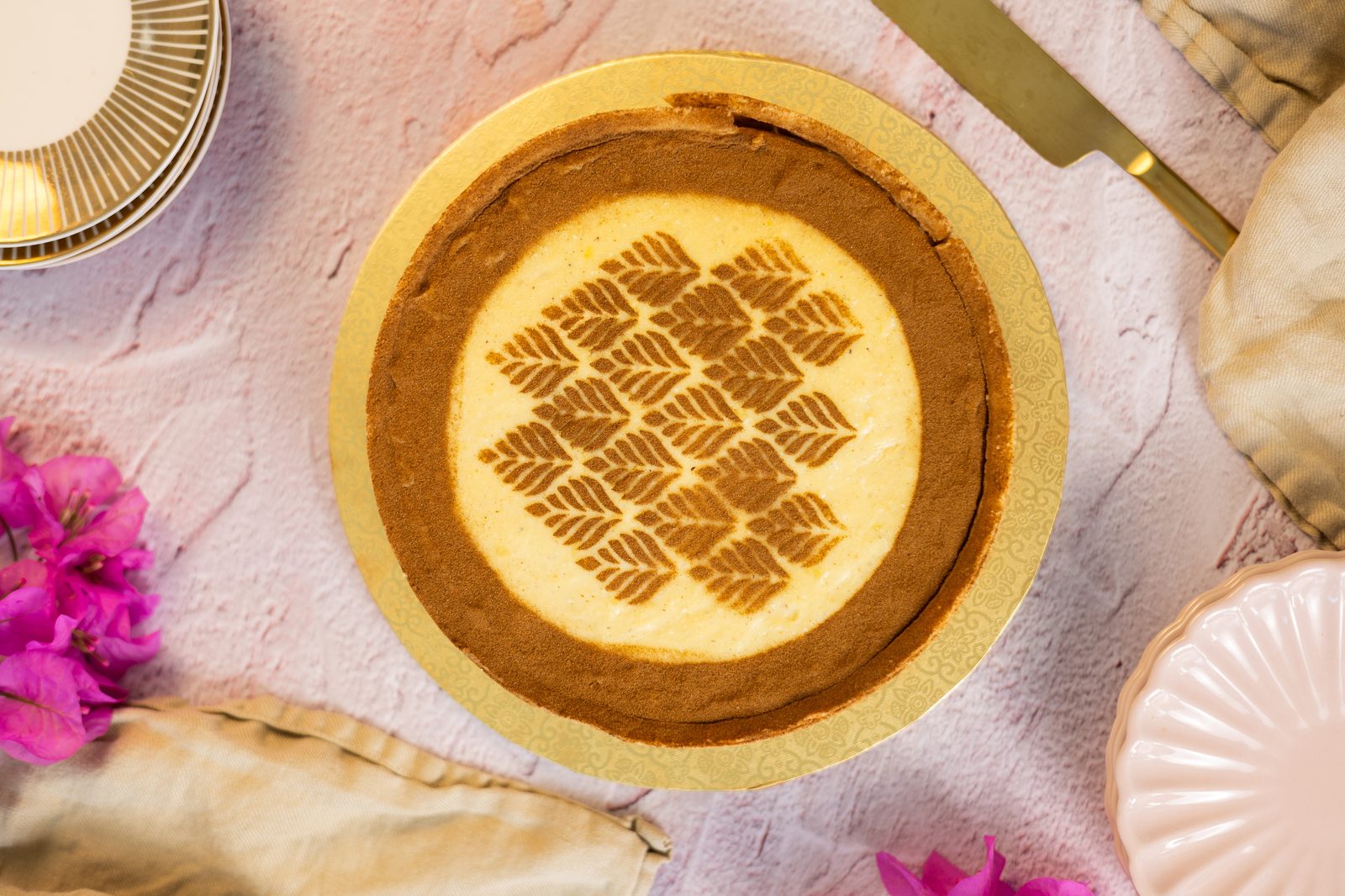 A whole no bake milk tart from above on a gold board, its cinnamon leaf stencil framed by flowers, plates and a cake server