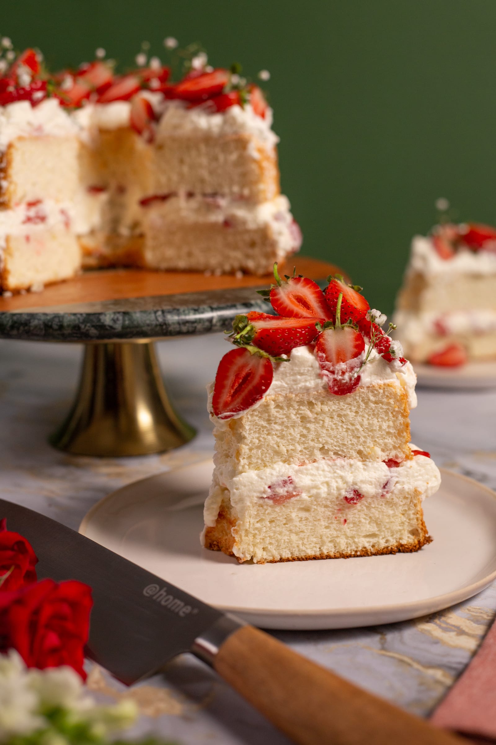 A slice of strawberry and cream angel food cake with the cut cake behind and a knife in front