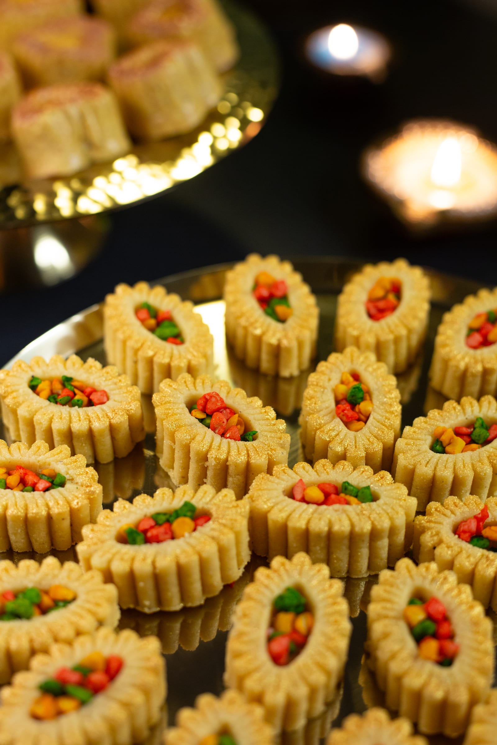 A gold plate piled with fresh cream burfee, with channa magaj on a stand behind