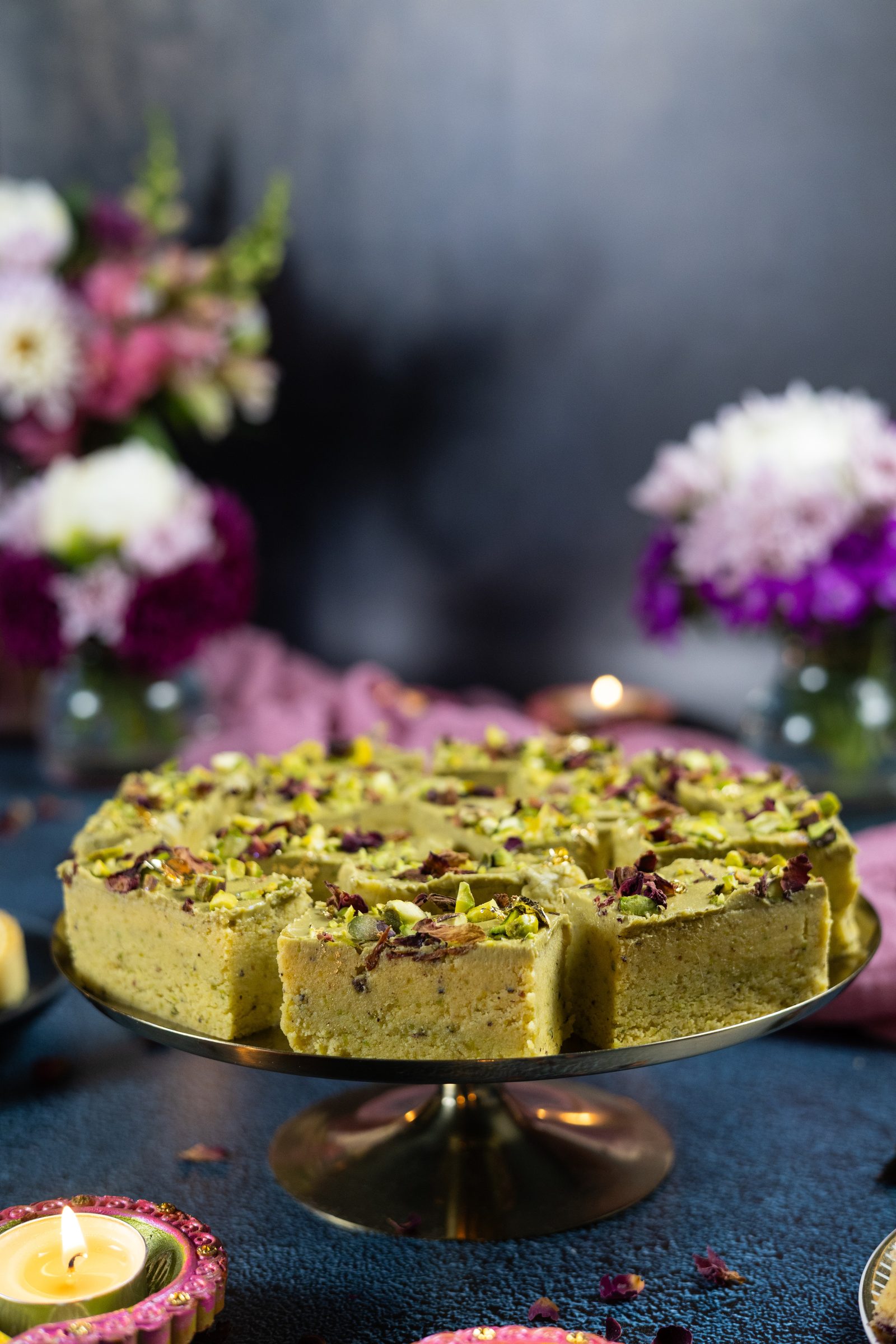 Pistachio burfee stacked on a gold cake stand, from the side, with flowers behind