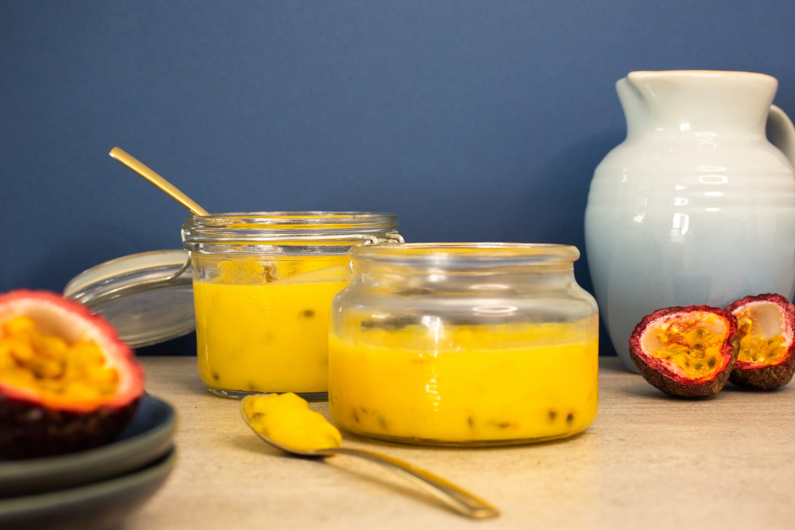 Two jars of passionfruit curd with a gold spoon, halved passionfruit and a blue jug