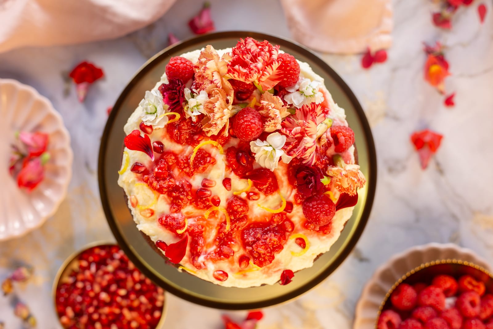 Lemon, rose and raspberry cake with white chocolate labneh ganache on a gold stand from above, with bowls of raspberries and pomegranate and rose petals on marble