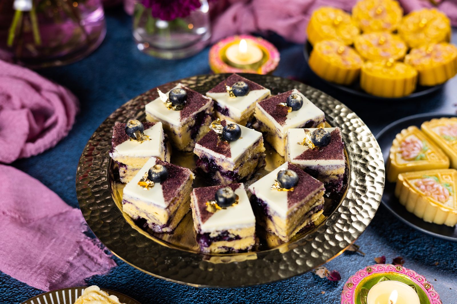 A hammered gold plate of blueberry ripple burfee topped with white chocolate and fresh blueberries, with mango burfee behind
