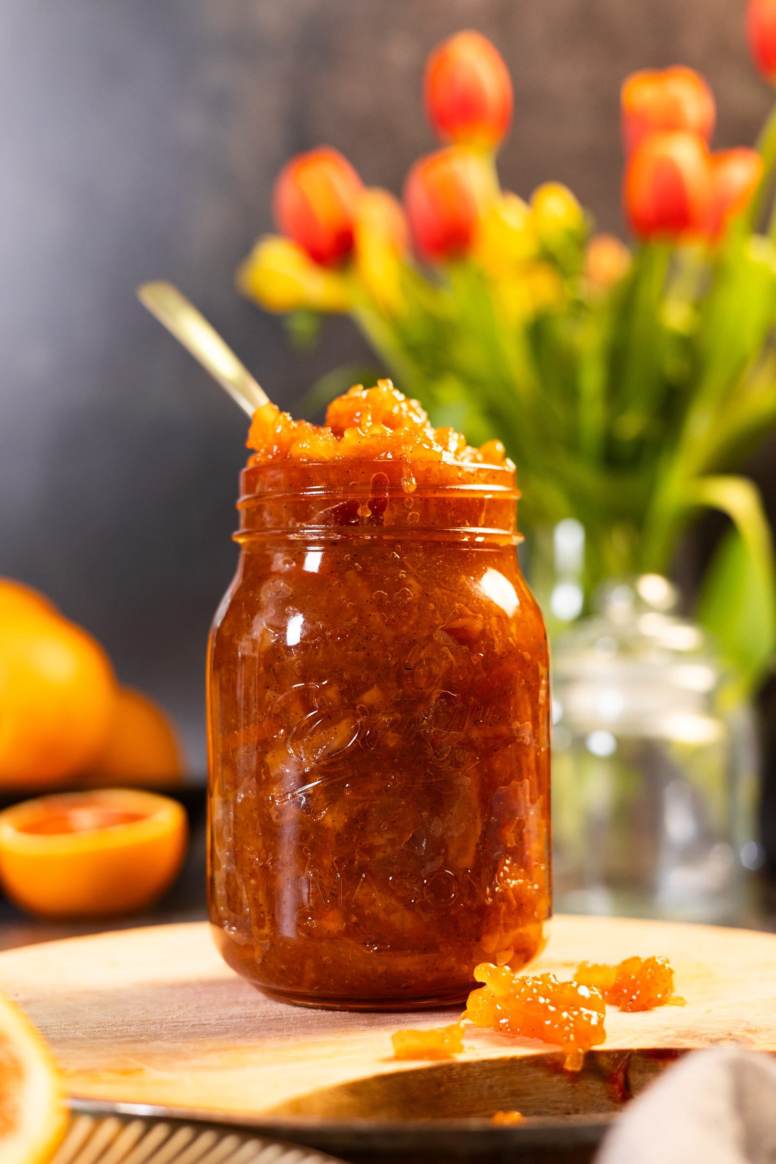 A jar of chunky blood orange marmalade with a spoon, in front of tulips