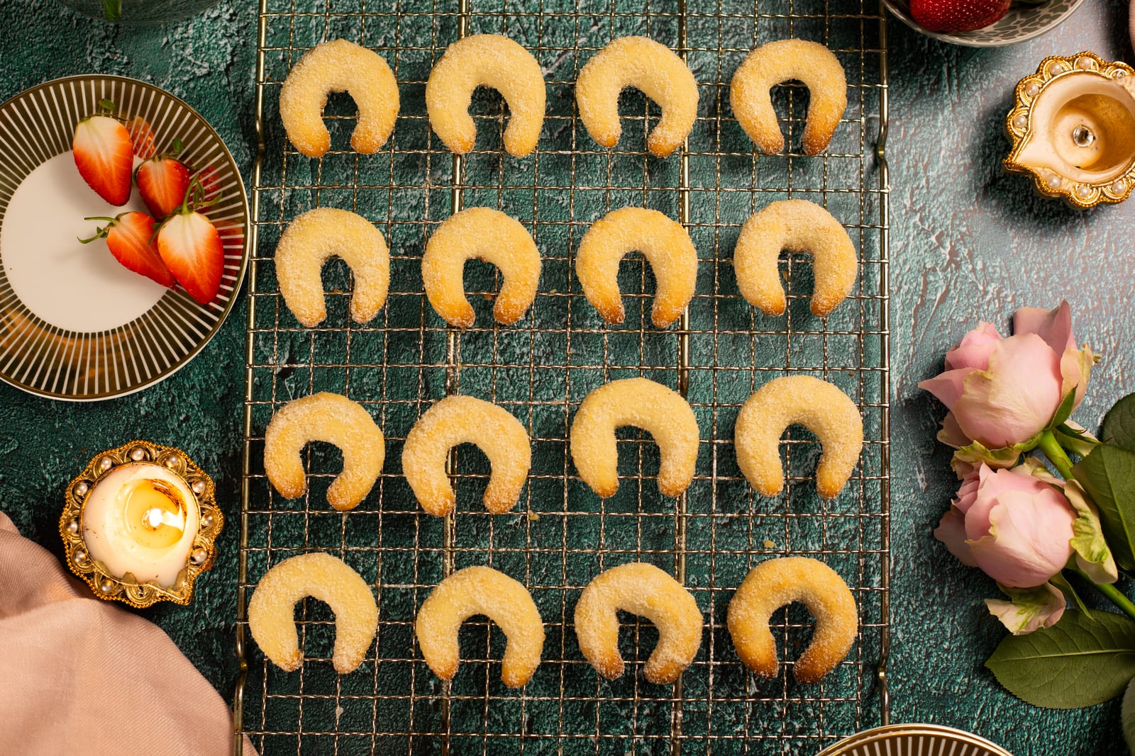 Rows of crescent-shaped cashew nut biscuits on a wire rack, from above, with diyas and roses