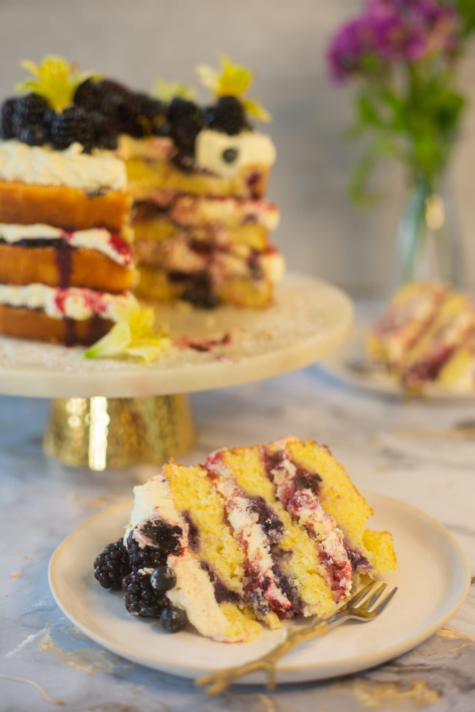 A slice of lemon and blueberry cake in front of the cut cake on its stand