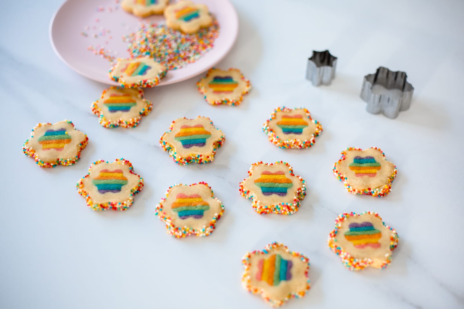 Rainbow cookies scattered on marble beside a pink plate of sprinkles and flower cutters