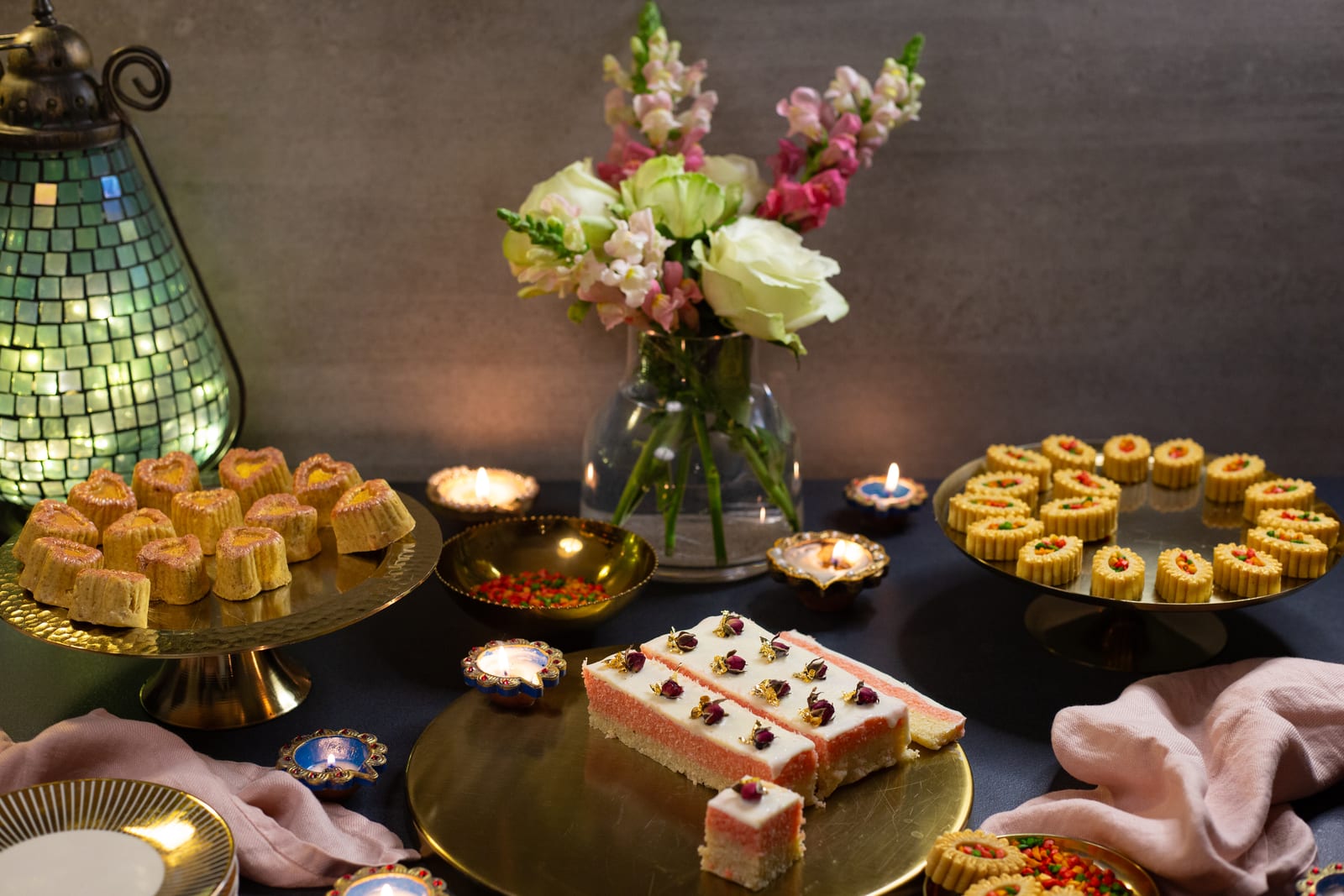 A Diwali table of Indian sweetmeats with fresh cream burfee, channa magaj and kopra paak among flowers, diyas and a mosaic lantern