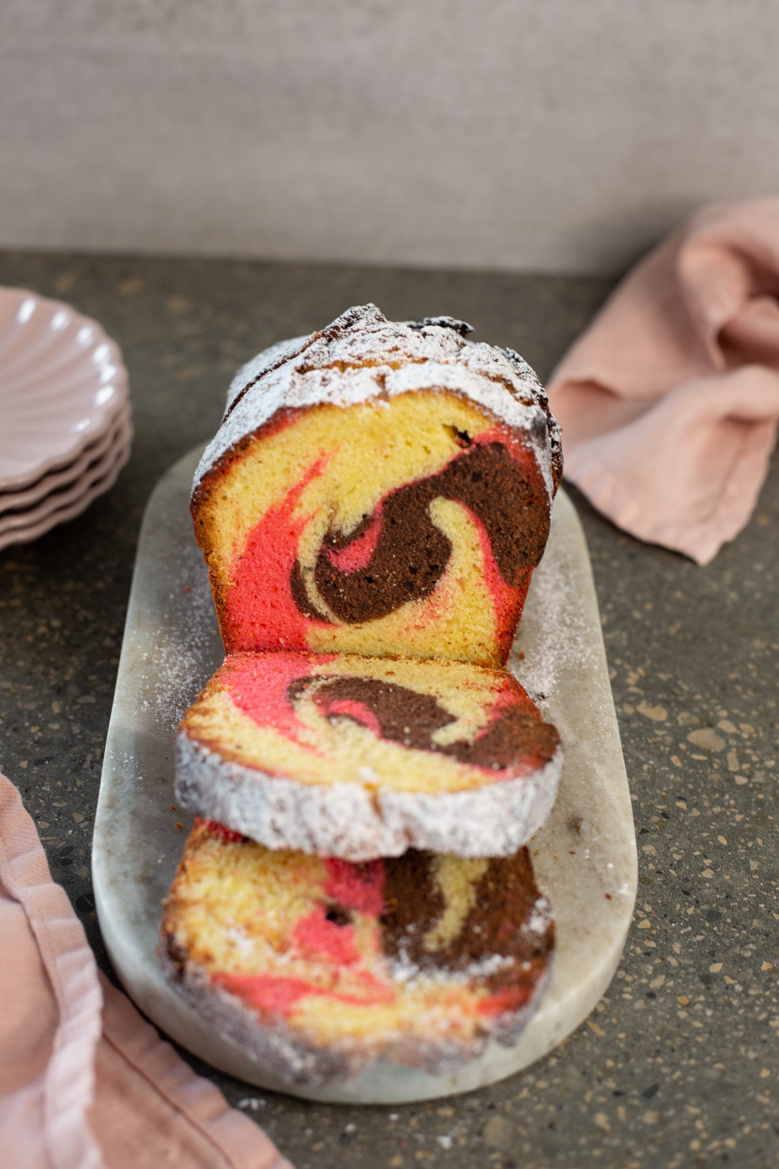A Neapolitan pound cake dusted with icing sugar, cut to show its vanilla, strawberry and chocolate swirl
