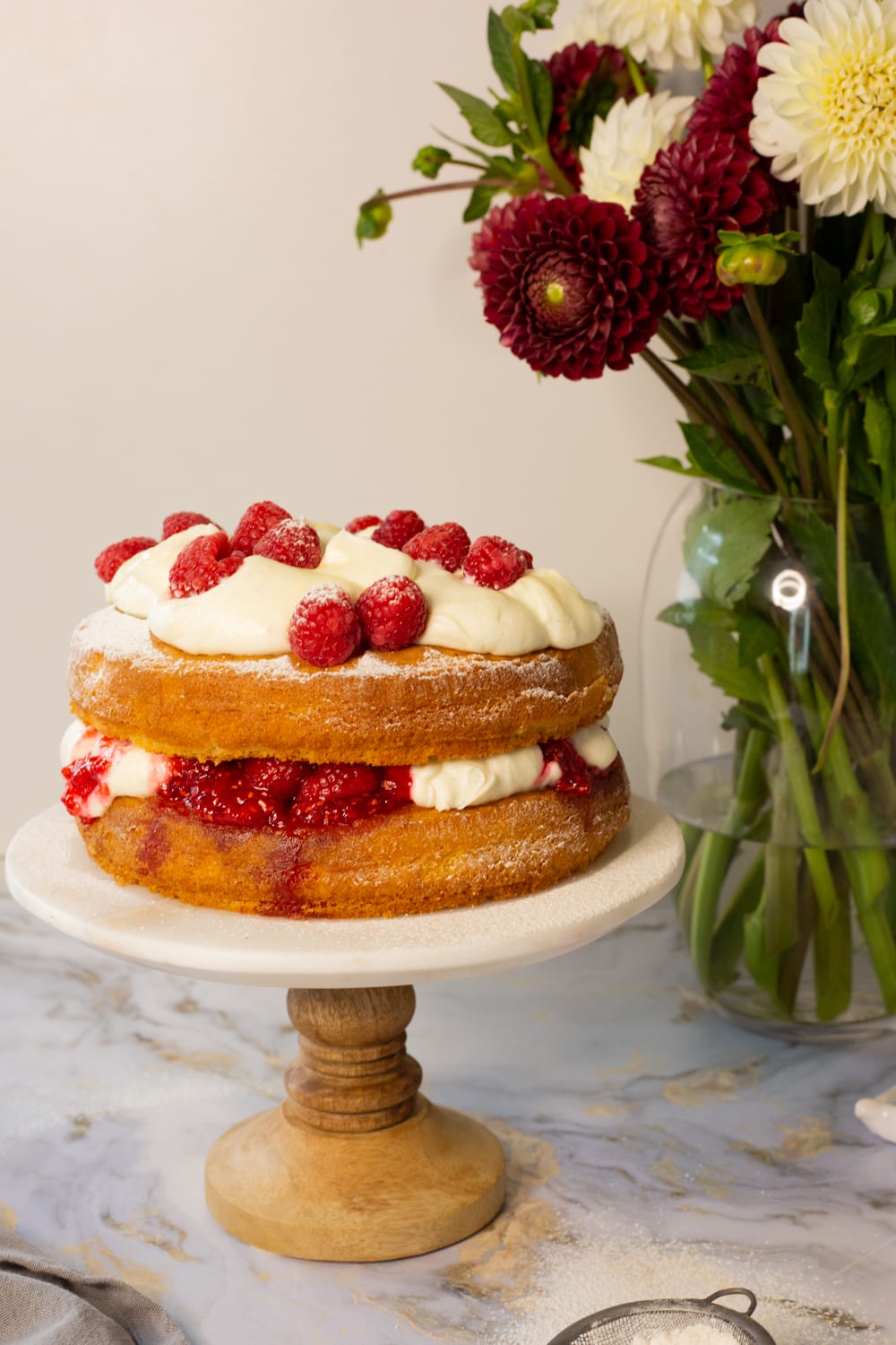 A whole Victoria sandwich topped with whipped cream and raspberries on a wooden cake stand, dahlias behind