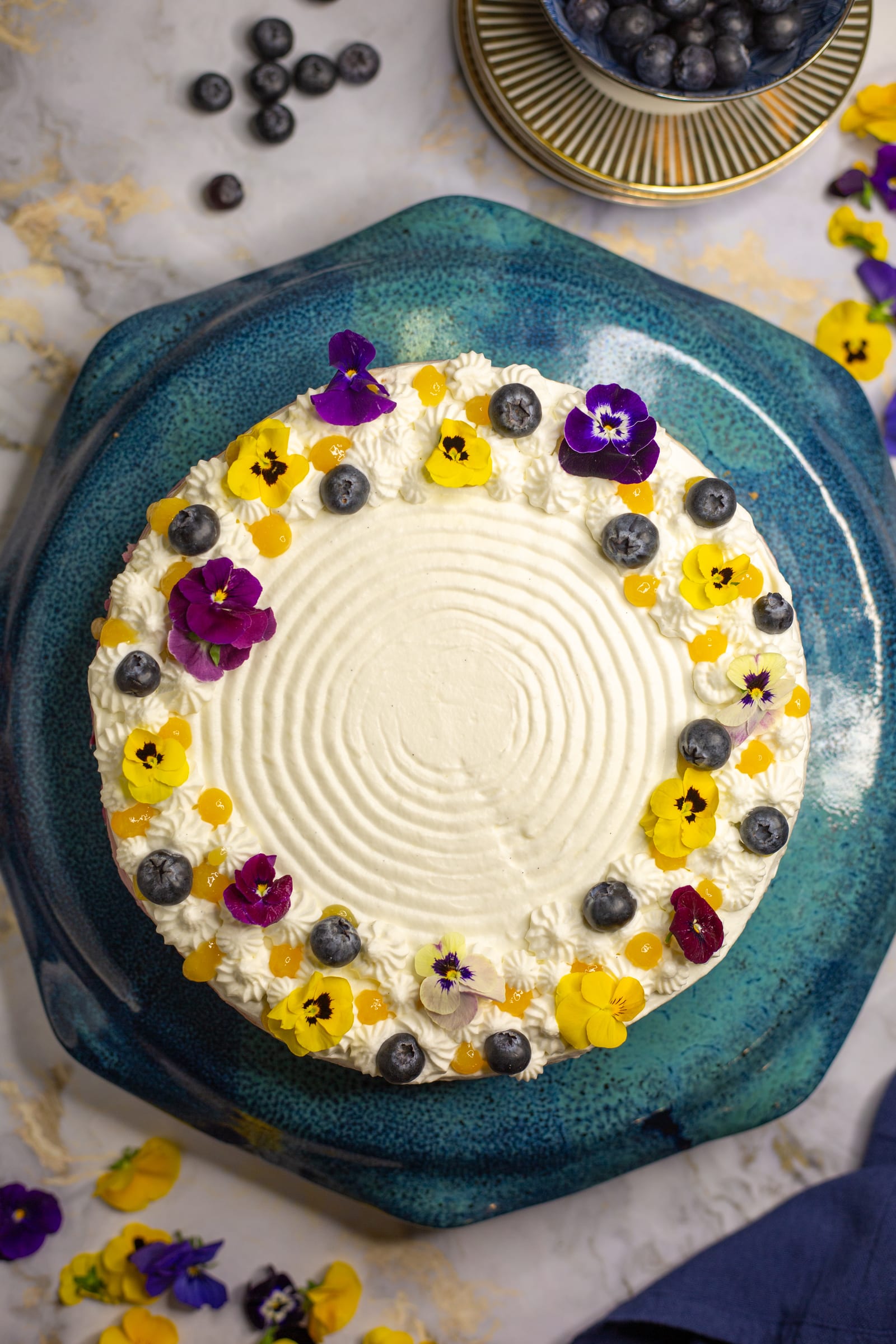 The lemon and blueberry cheesecake from above on its teal stand, crowned with pansies and blueberries