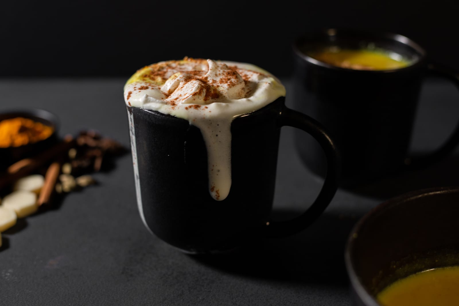 A mug of turmeric and ginger white hot chocolate with whipped cream dripping down its side, spices and white chocolate beside it