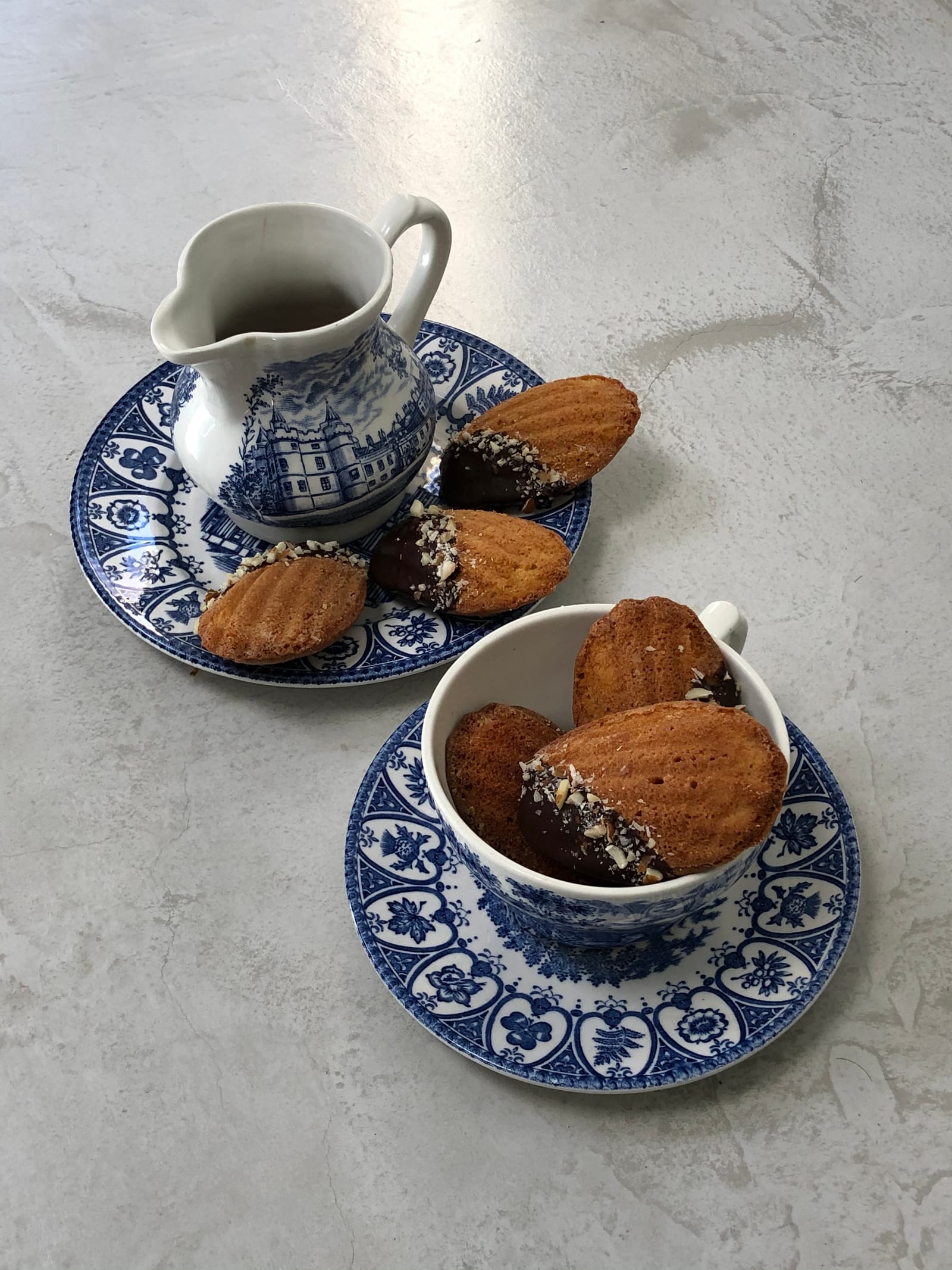A teacup of brown butter and almond madeleines beside a blue and white jug and saucer, from above