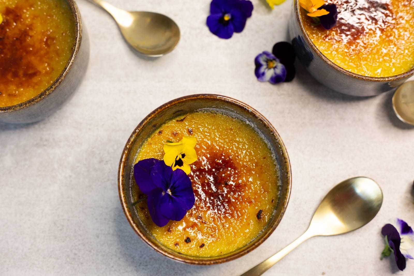 A chai-spiced crème brûlée topped with a purple viola beside gold spoons and more ramekins, from above