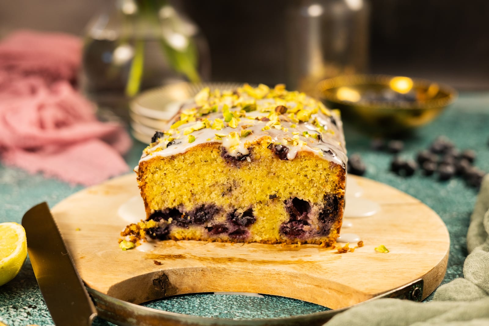 A pistachio, blueberry and lime loaf cut open on a wooden board, with flowers and jars behind