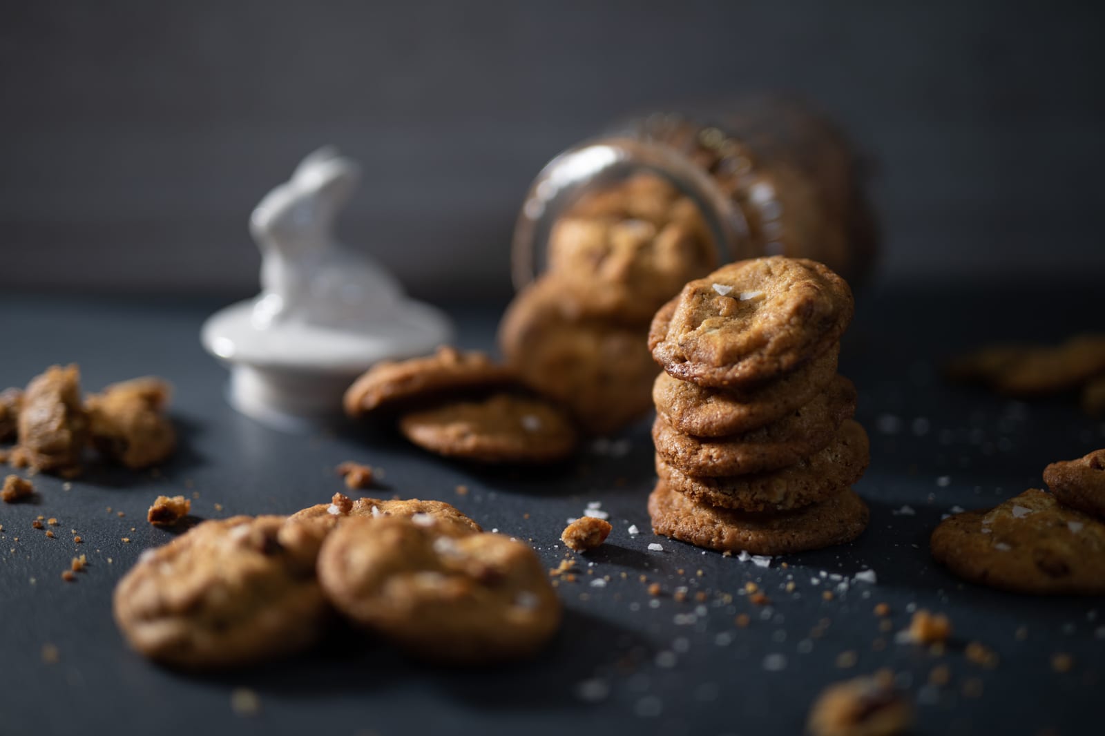 A jar with a bunny lid tipped over, spilling pecan and white chocolate salted cookies, a stack of cookies beside it