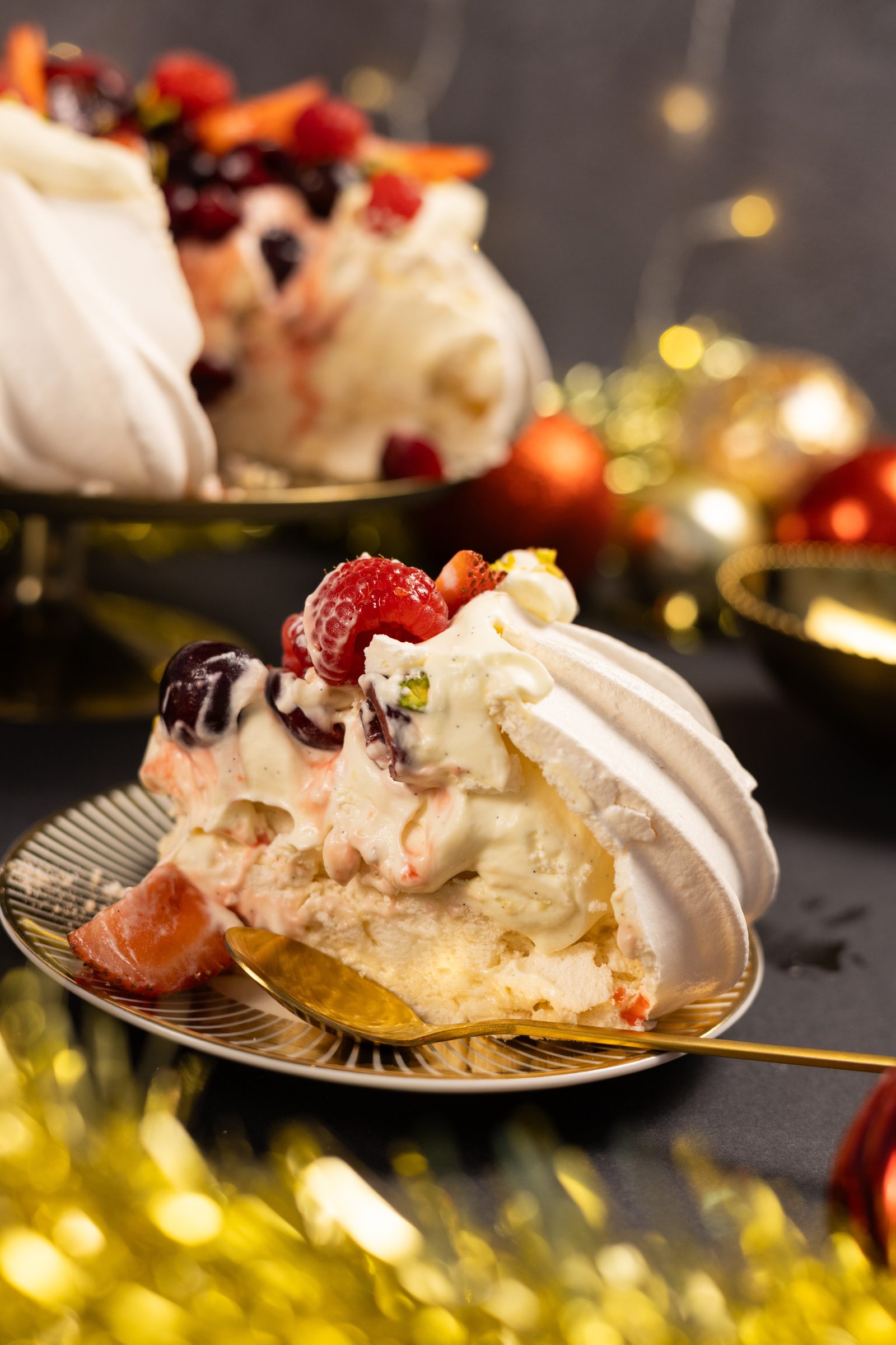 A slice of very berry pavlova on a gold plate with a fork, the cut pavlova behind