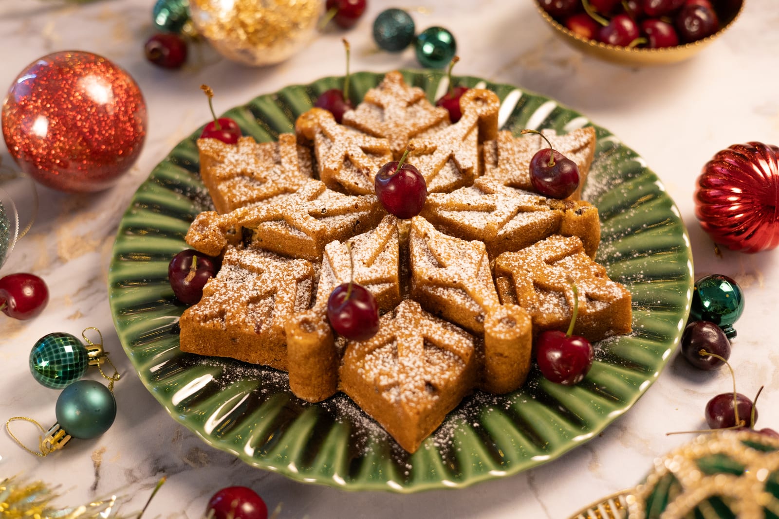 A snowflake-shaped eggless fruitcake dusted with icing sugar and topped with cherries, among Christmas baubles