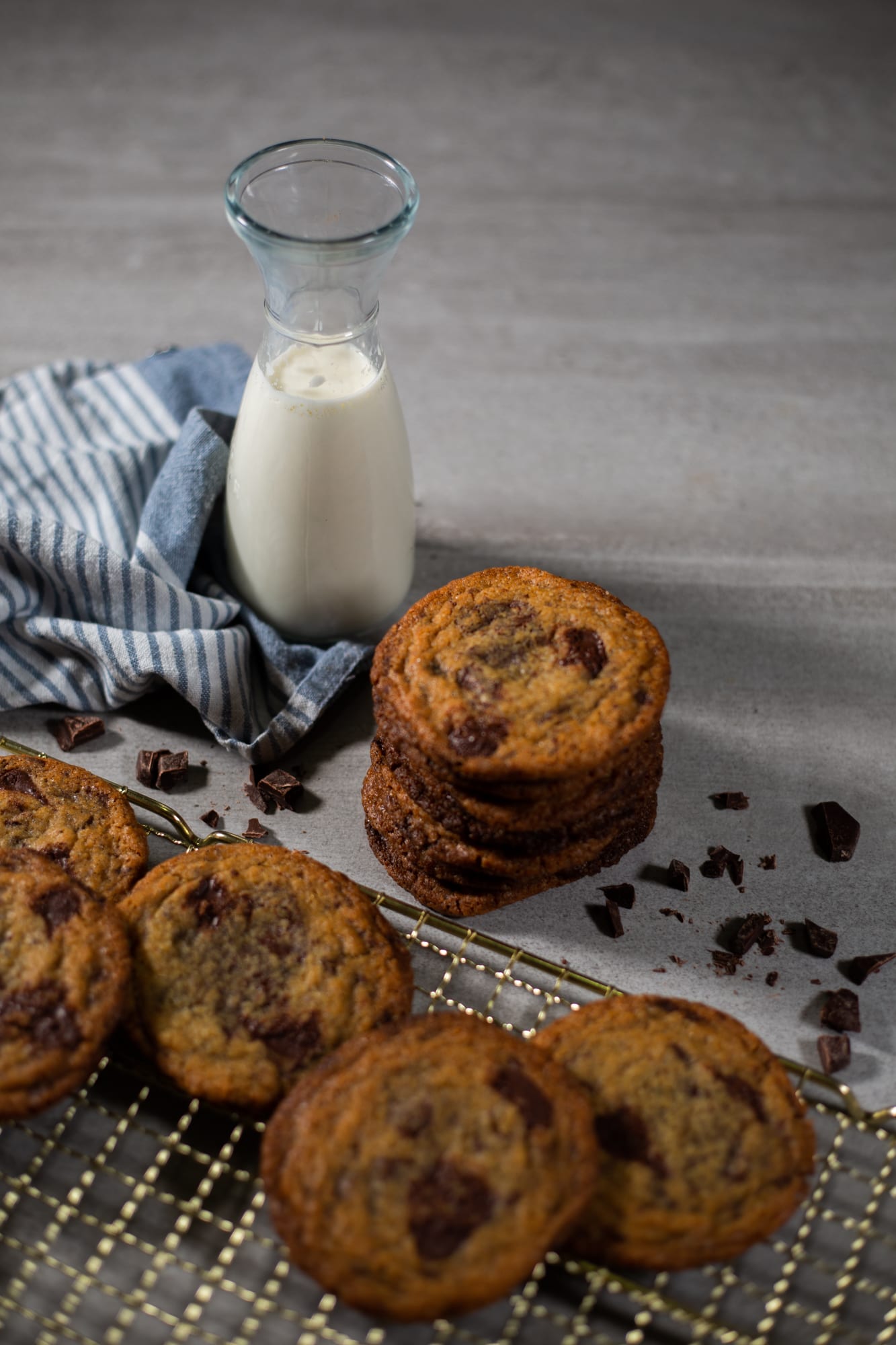 A stack of pan banging chocolate chip cookies on a gold rack beside a milk carafe
