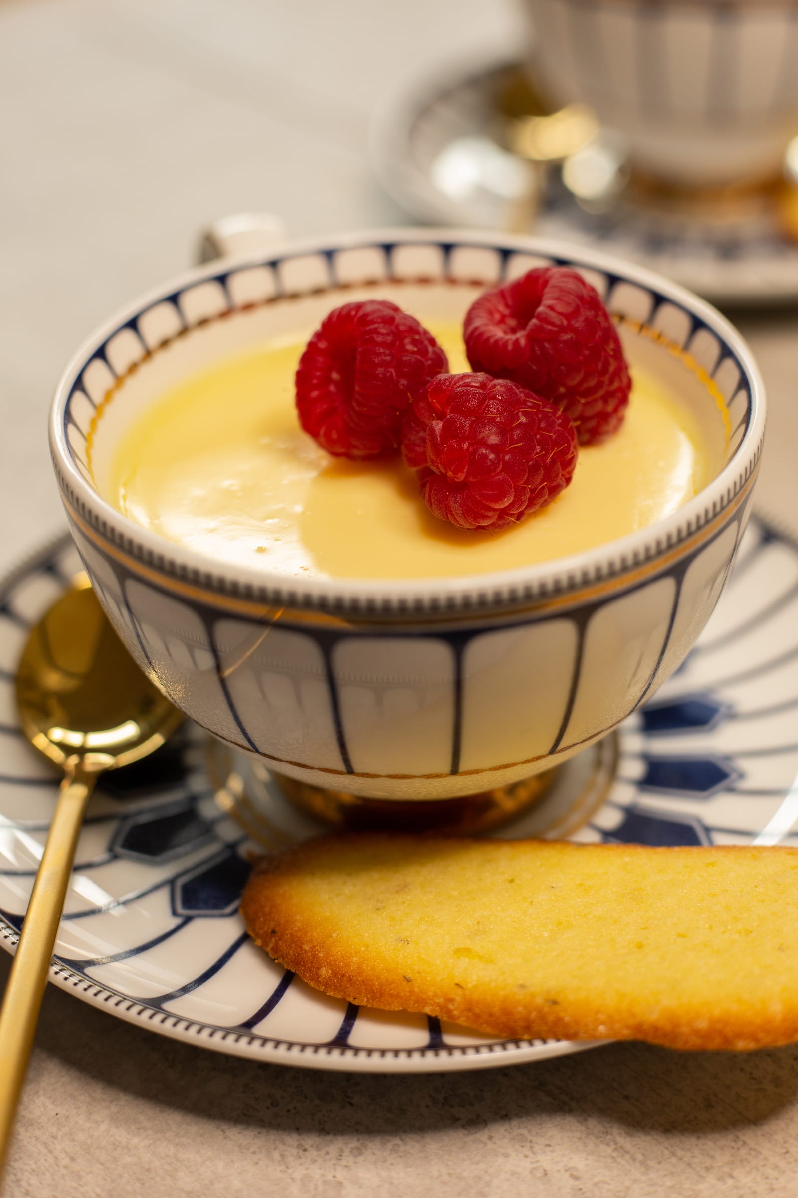 A lemon posset in a teacup from the side, with raspberries and a cat's tongue biscuit