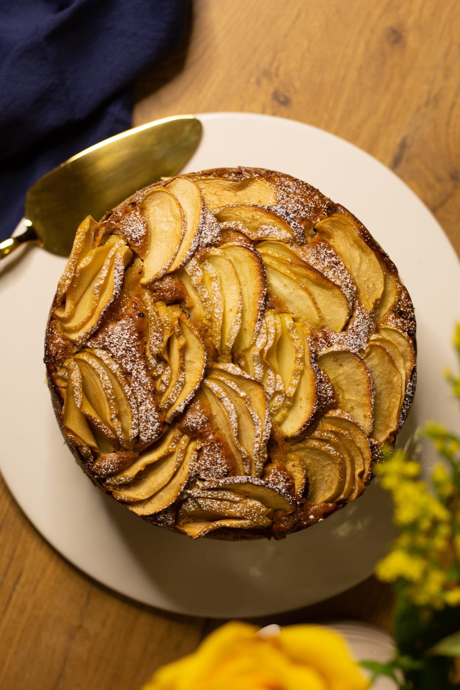 A whole apple and olive oil cake with a gold cake server and yellow roses, from above