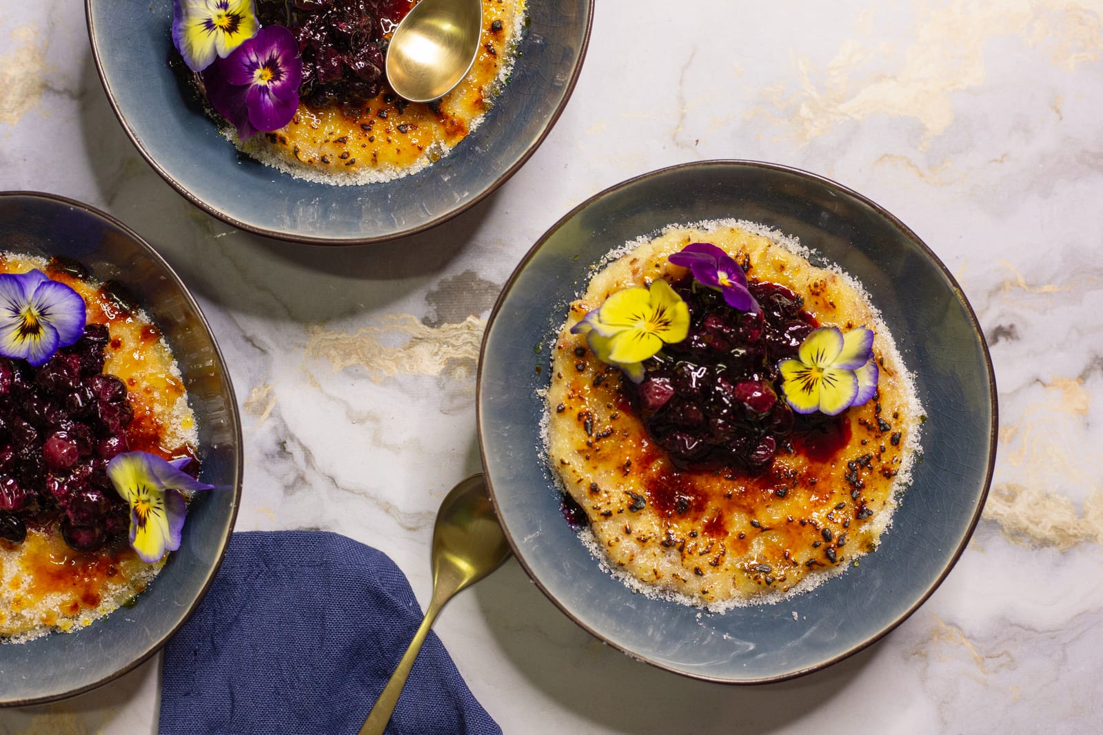 Bowls of kheer brûlée topped with blueberry compote and violas on marble, with a gold spoon and navy napkin, from above