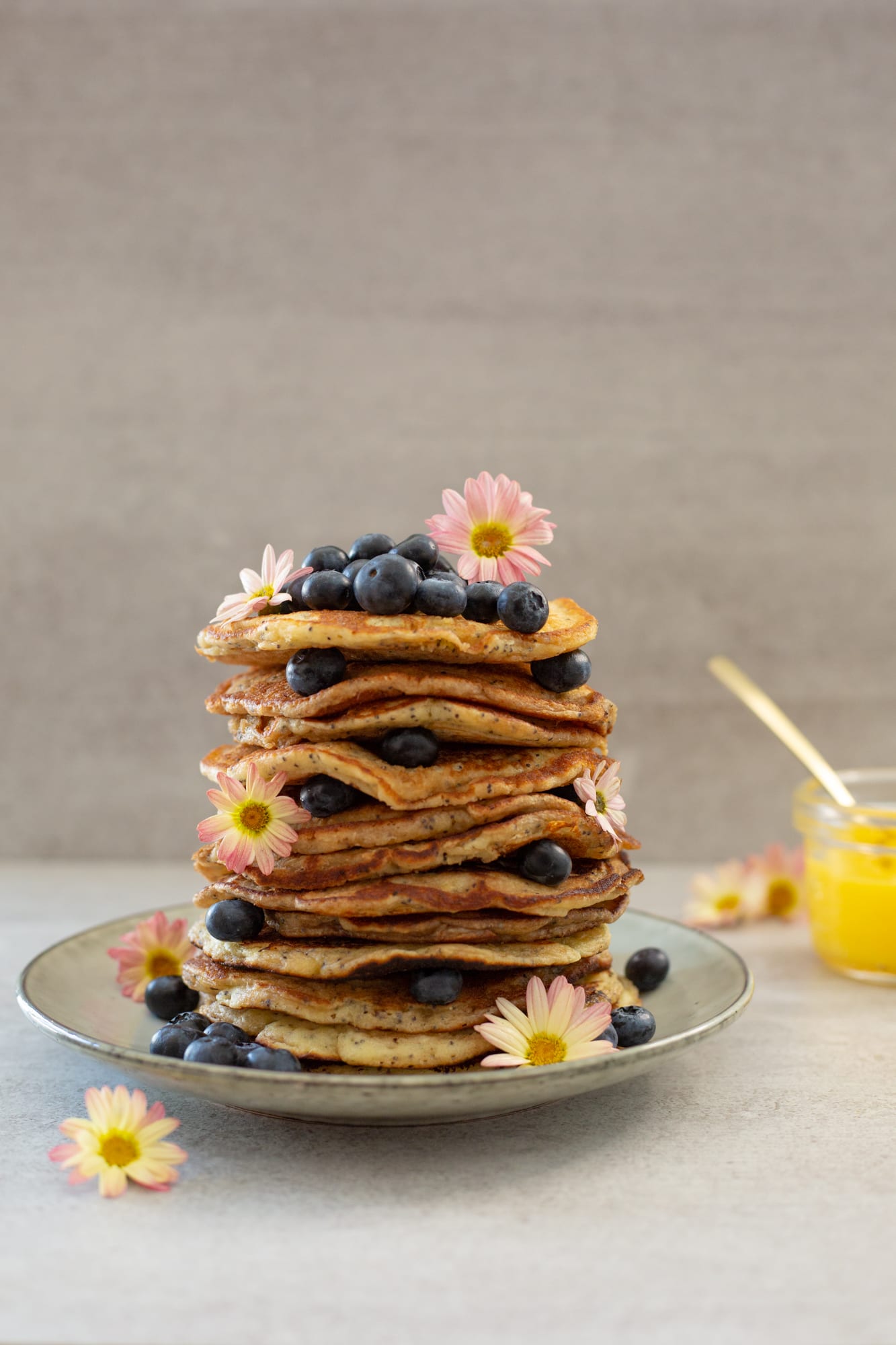 A stack of lemon and poppyseed pancakes with blueberries and small pink flowers, before the curd goes on