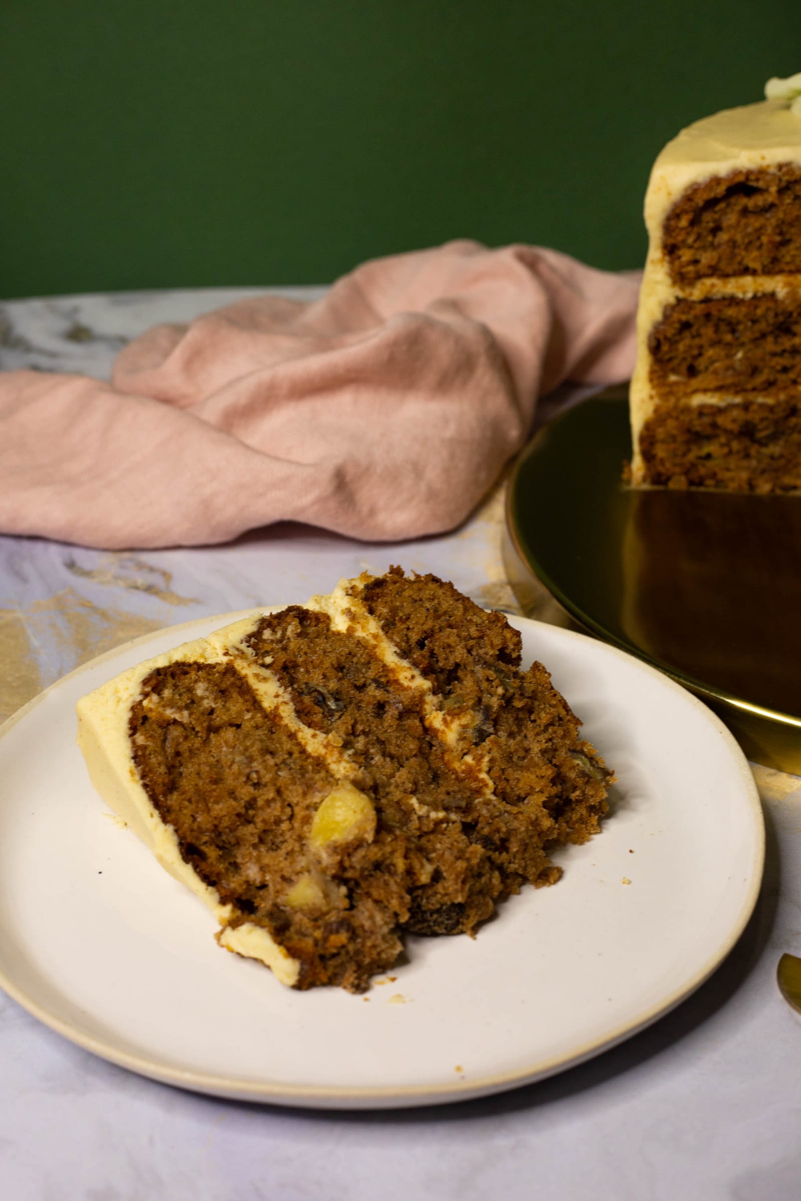 A slice of hummingbird cake on a white plate in front of the cake, against a green wall